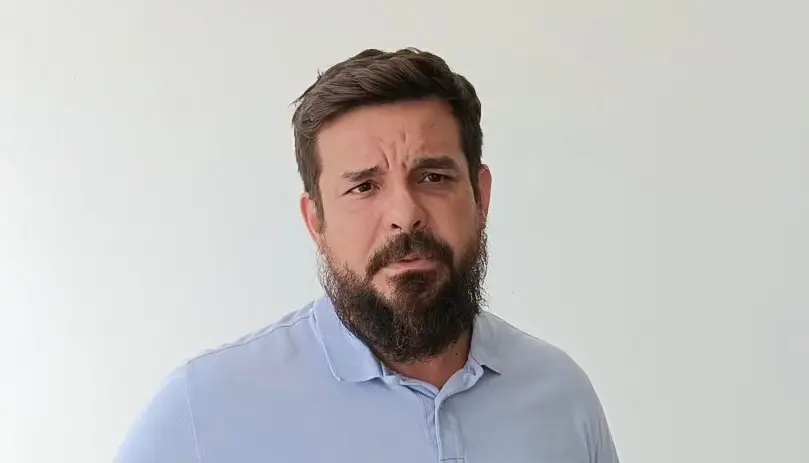 Foto horizontal que mostra o juiz Thalles Nóbrega, do busto pra cima, em frente a uma parede branca. Ele é um homem branco, de cabelo, barba e olhos castanhos, usando camisa social azul clara.