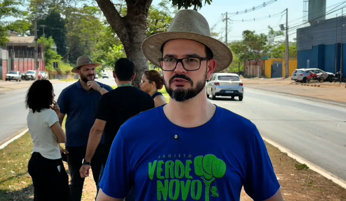 Homem de barba, óculos de armação preta e chapéu de palha, veste camiseta azul do Projeto Verde Novo e é entrevistado em avenida; ao fundo, pessoas conversam.