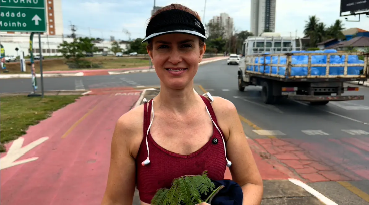 Mulher de viseira preta e top vinho, com fones de ouvido no pescoço, segura folhas de uma muda e é entrevistada em ciclovia vermelha de avenida.