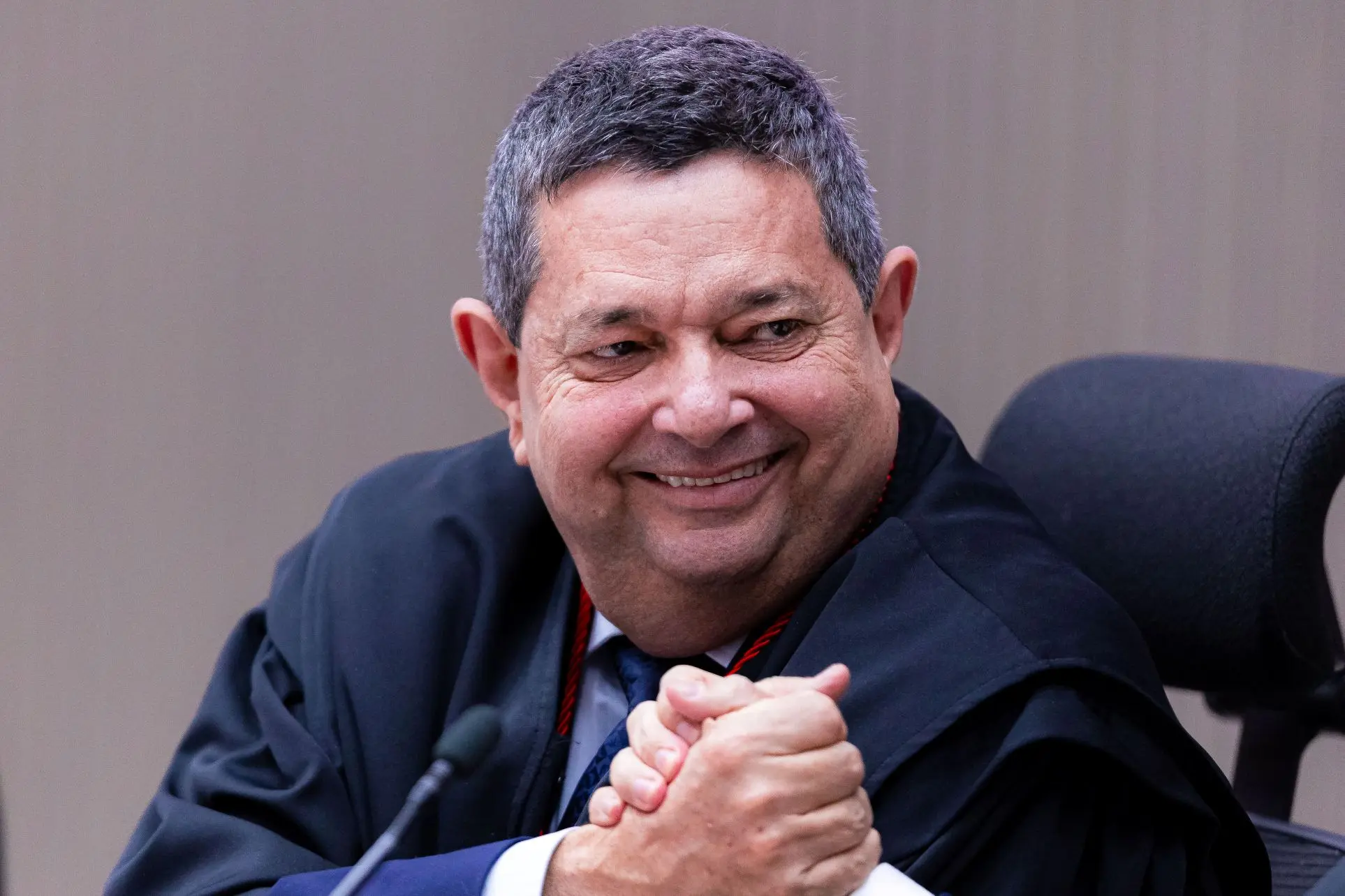 Foto horizontal em plano fechado do desembargador José Luiz Leite Lindote, sentado, olhando sorrindo para o lado e apertando a mão de outro homem. Ele é um homem branco, de cabelos grisalhos, usando toga.