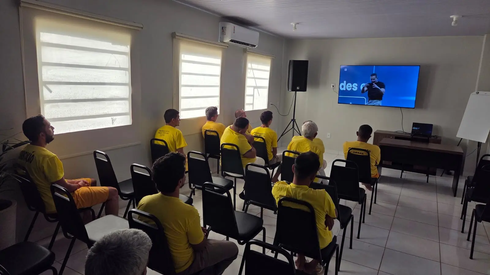 Homens de uniforme amarelo estão sentados em cadeiras pretas e assistem um vídeo com um homem gesticulando, em sala com ar-condicionado.