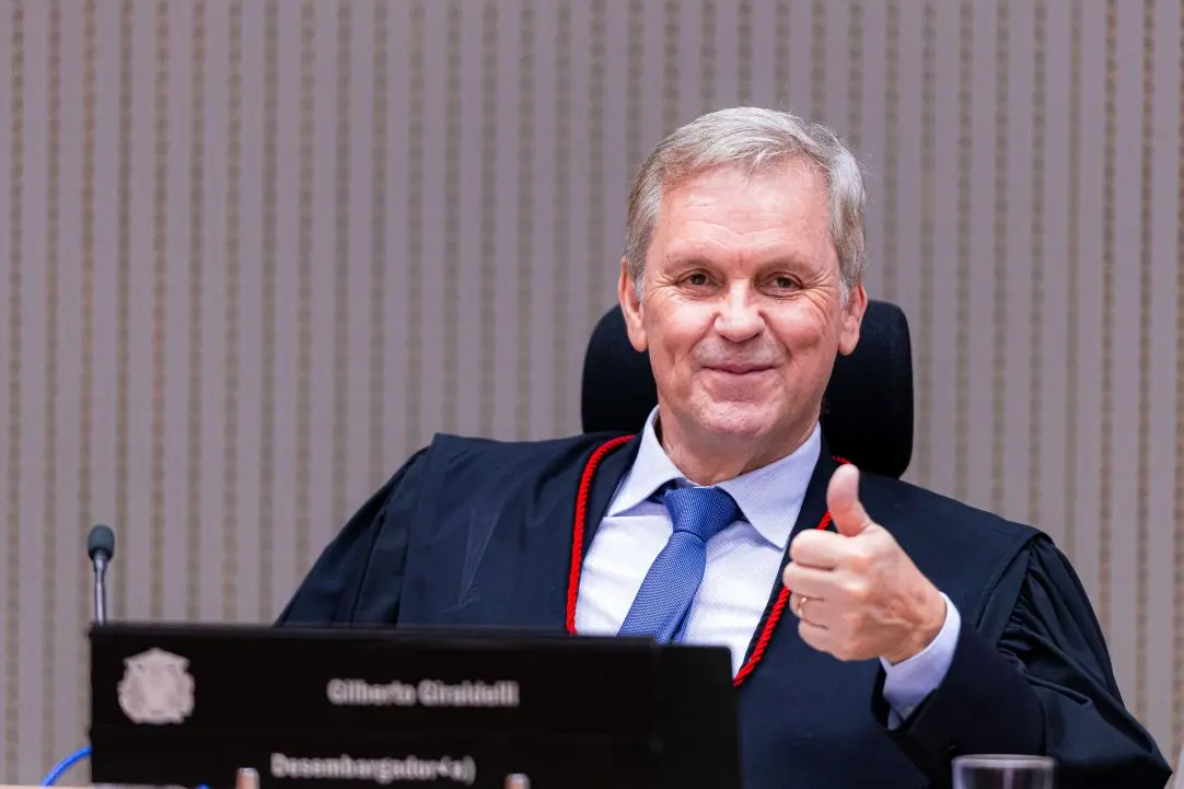 Foto horizontal que mostra o desembargador Gilberto Giraldelli sorrindo e fazendo sinal de joia com a mão esquerda, sentado em sua poltrona no Plenário do TJMT. Ele é um homem branco de cabelos brancos, olhos claros, usando toga. 