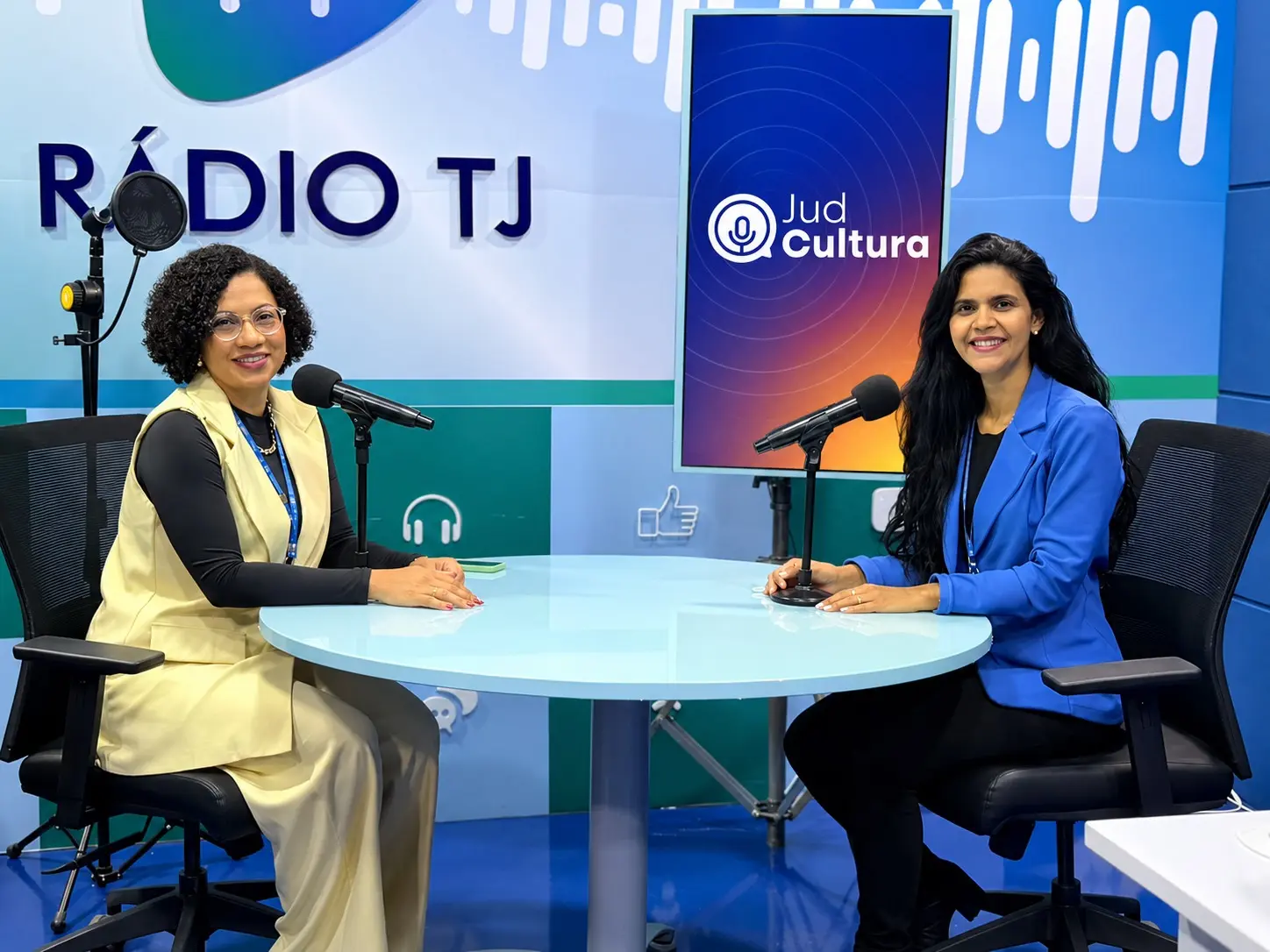 Duas mulheres sorriem, sentadas à mesa do estúdio da Rádio TJ, diante de microfones e do painel do programa Jud Cultura. Uma veste colete amarelo-claro; a outra, blazer azul.