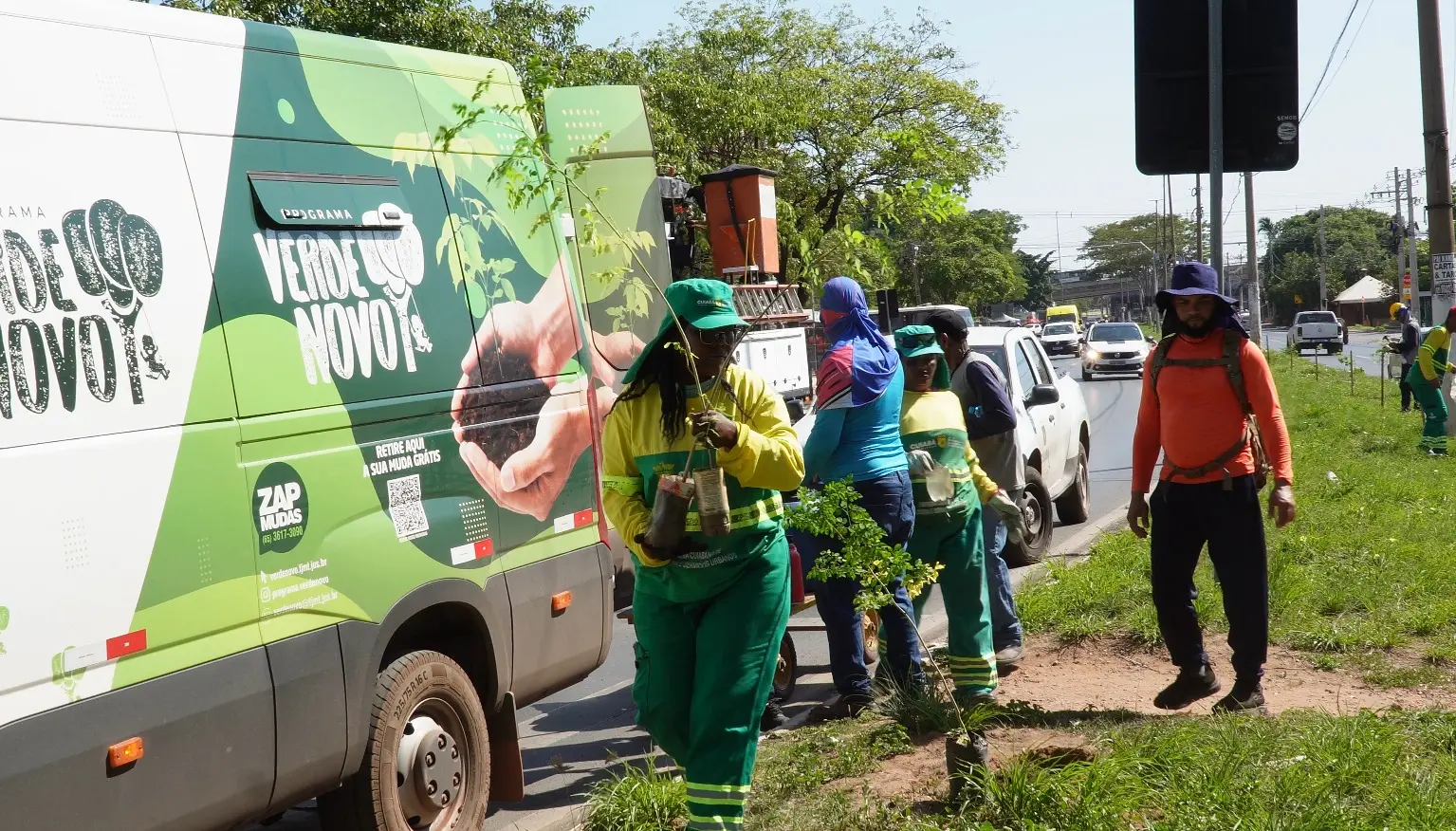 Agentes da Limpurb, de uniforme verde e amarelo, carregam mudas ao lado de van do Programa Verde Novo, em canteiro de avenida movimentada.