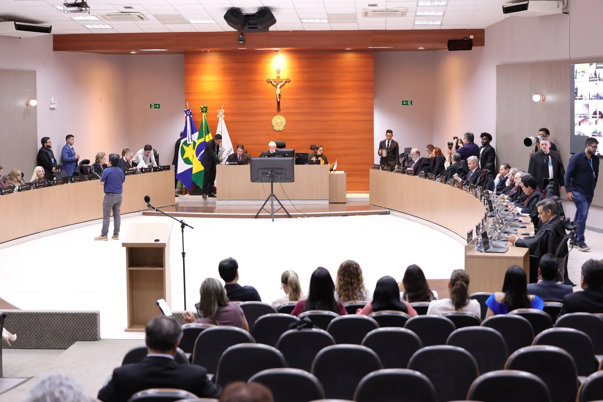 Foto horizontal em plano aberto que mostra o plenário 1 do TJMT, vista do fundo, com pessoas sentadas nas poltronas e os desembargadores sentados em volta da grande mesa oval. 