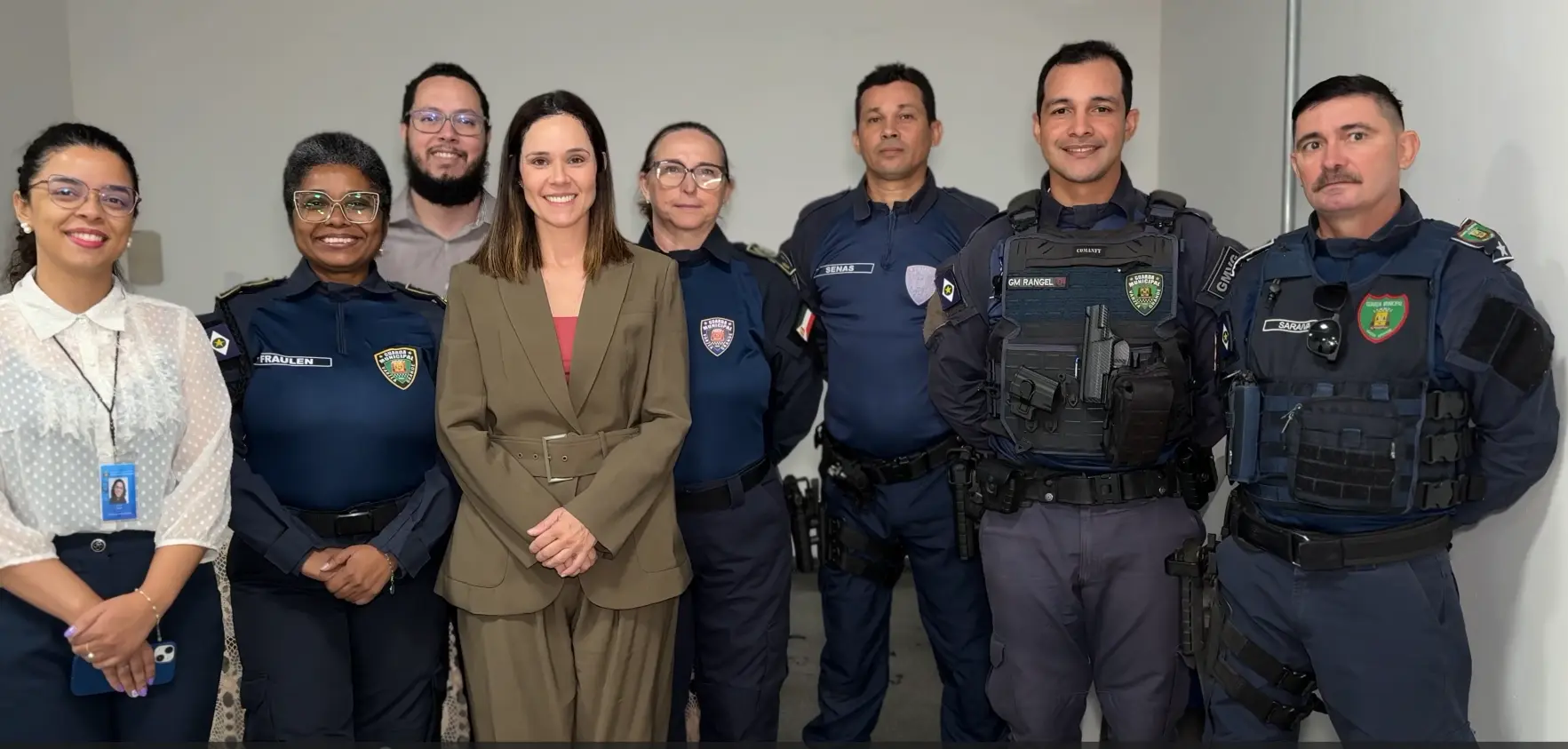 Sete pessoas posam lado a lado sorrindo. Entre elas, civis e integrantes da guarda municipal com fardamento azul-escuro e coletes táticos. O fundo é uma parede clara e lisa.