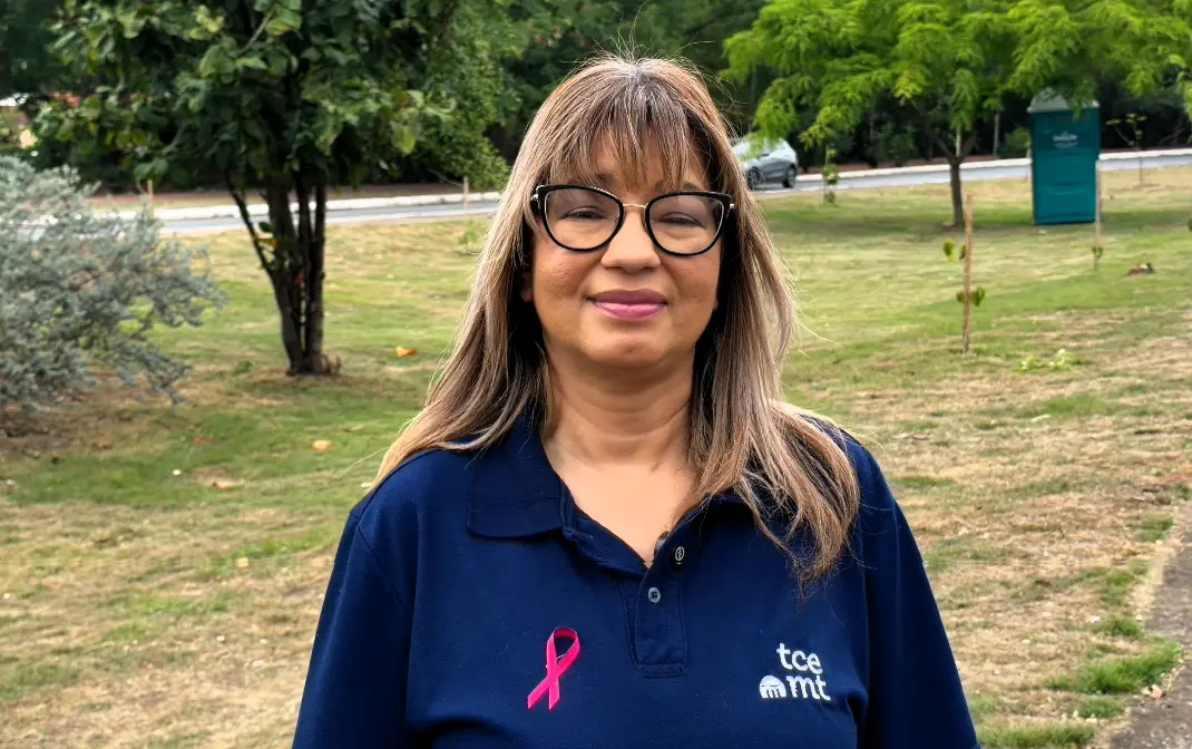 Mulher de cabelos longos e claros, franja e óculos de armação preta, veste polo azul-marinho do TCE-MT com laço rosa e é entrevistada em área verde.