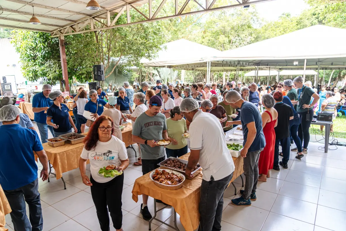 Pessoas servem-se em mesas de buffet cobertas com toalhas amarelas em área coberta ao ar livre. Funcionários de touca auxiliam no atendimento em evento movimentado e arborizado.