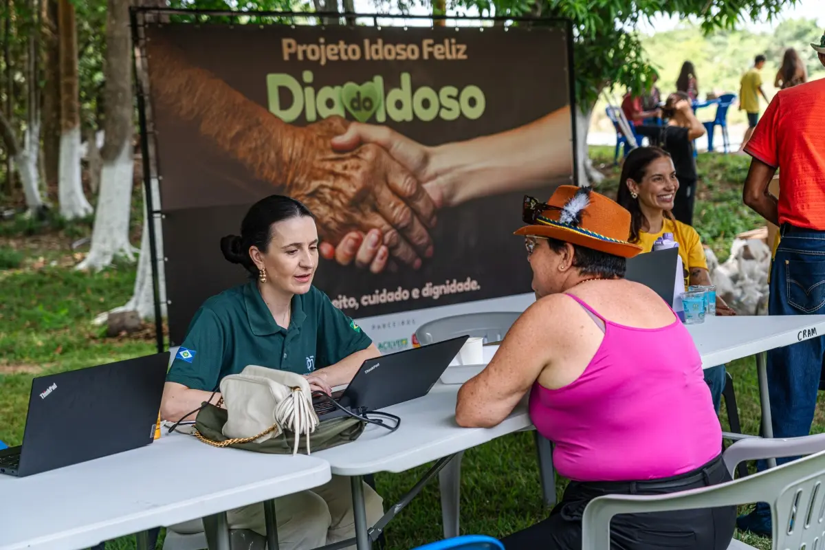Atendente de camisa verde atende mulher de regata rosa e chapéu em mesa ao ar livre com notebook. Ao fundo, banner do "Projeto Idoso Feliz - Dia do Idoso" no gramado.