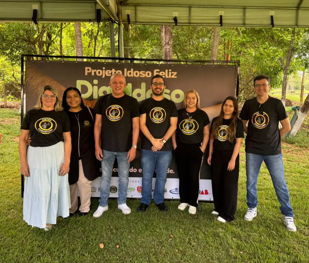 Sete pessoas posam sorrindo em gramado sob tenda. Seis usam camisetas pretas com logo circular dourado. Ao fundo, banner do "Projeto Idoso Feliz - Dia do Idoso" com logos de apoiadores abaixo.