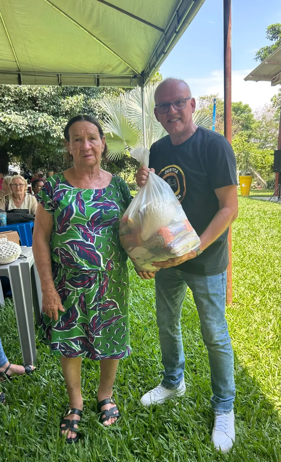 Homem sorridente de camiseta preta e jeans entrega sacola com alimentos a uma mulher de vestido estampado verde. Estão em pé sobre o gramado sob a sombra de uma tenda.