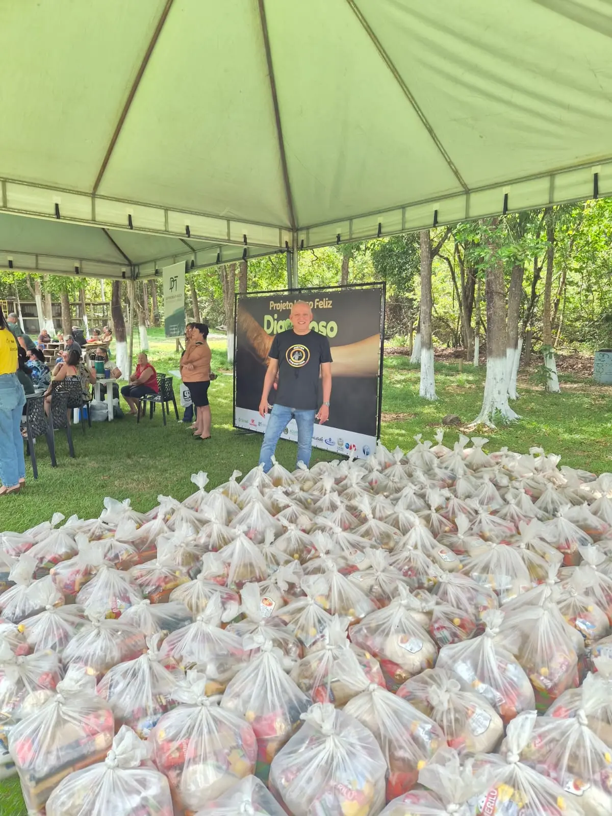 Homem em pé sob tenda ao ar livre com dezenas de sacolas de cestas básicas organizadas no gramado à sua frente. Ao fundo, banner do "Projeto Idoso Feliz - Dia do Idoso" e área verde.