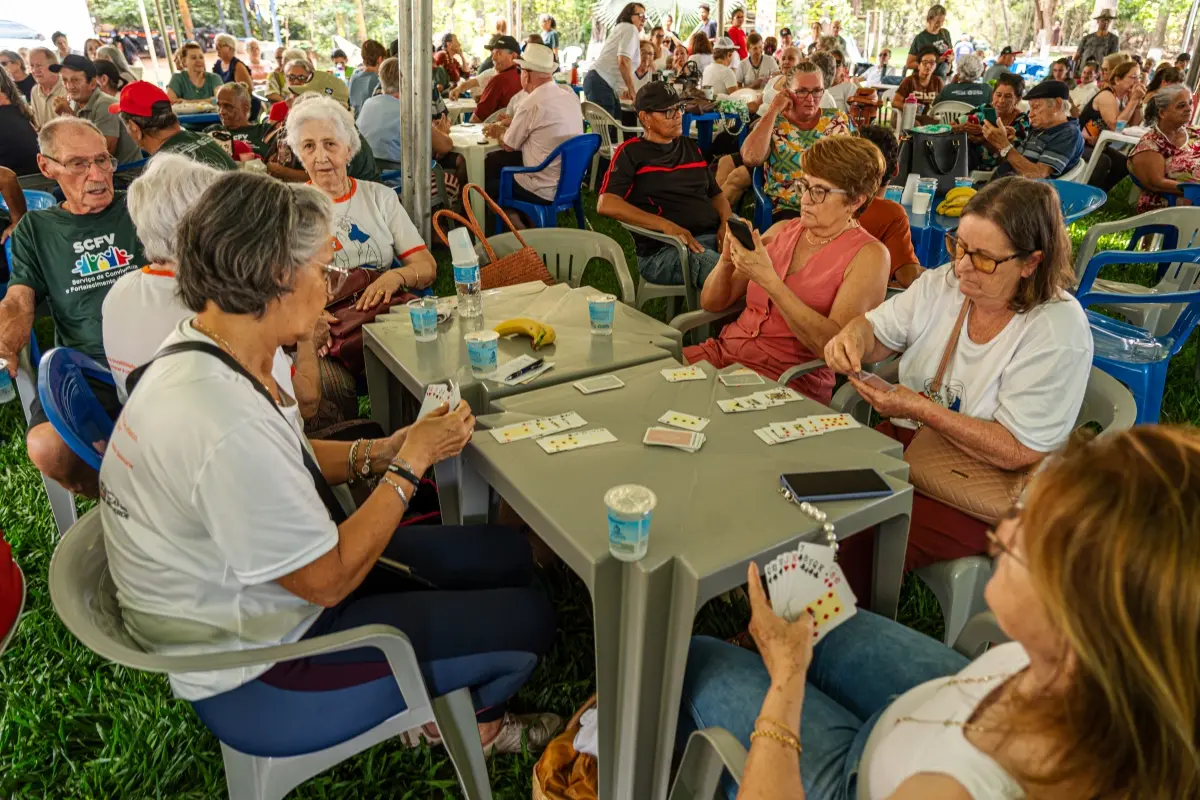 Grupo de idosos joga baralho em mesas cinzas dispostas sobre o gramado sob tenda coberta. Ao fundo, muitas pessoas conversam em espaço de convivência ao ar livre.