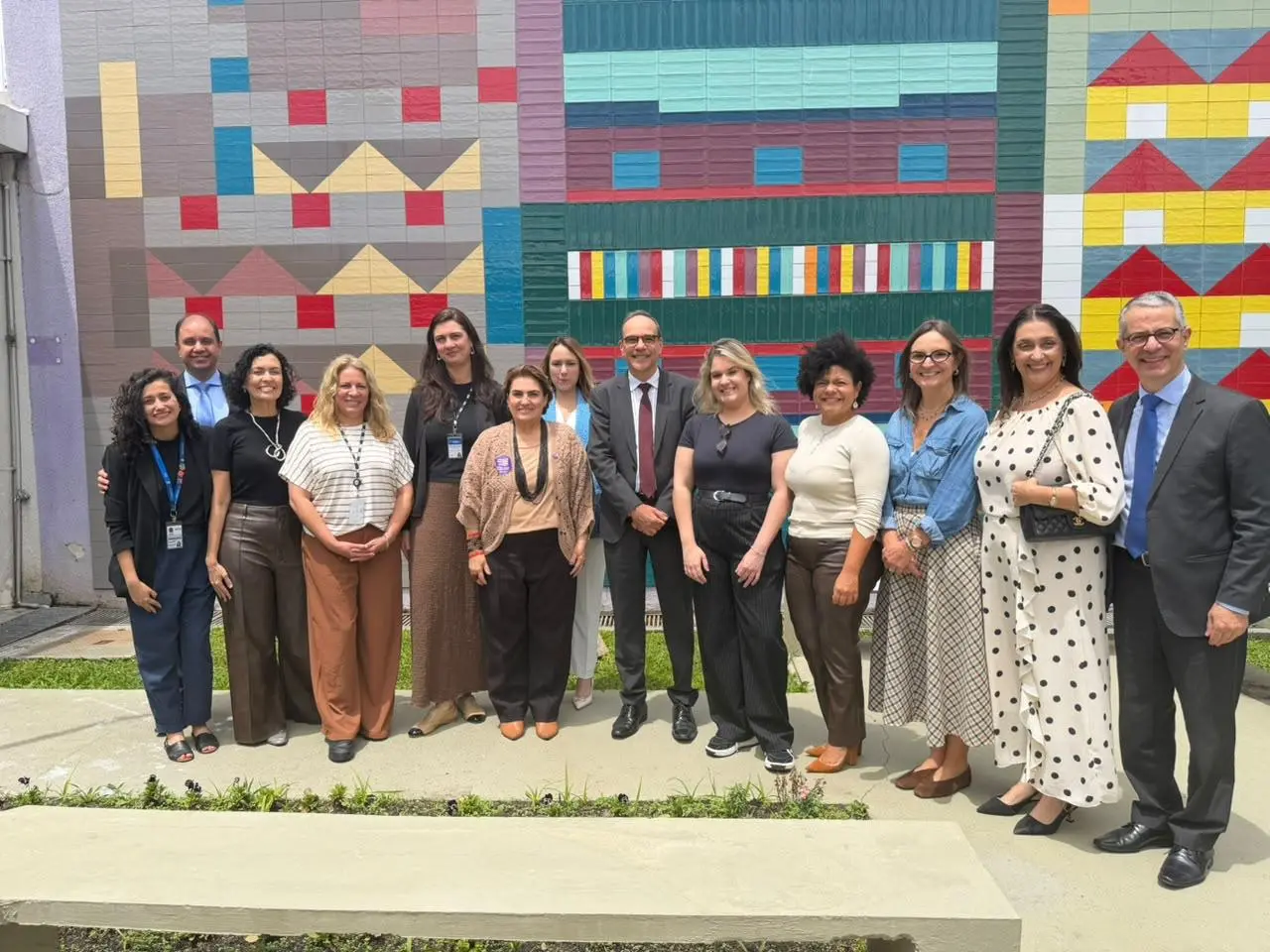 Grupo de 13 pessoas posa em pátio ao ar livre, diante de painel de azulejos coloridos com formas geométricas. Alguns usam crachás, e há homens de terno e mulheres em trajes casuais.