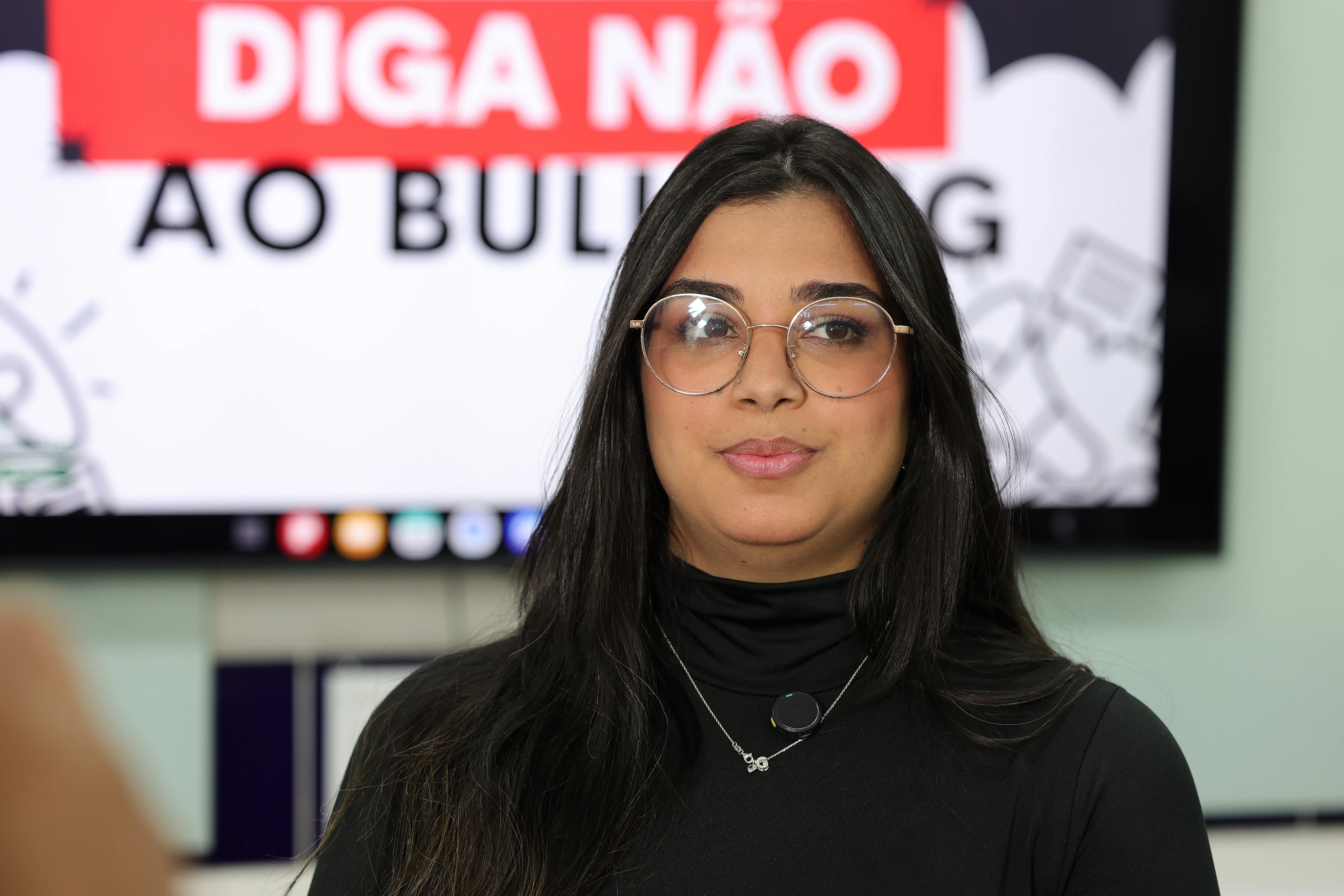 Retrato em plano médio de uma mulher de óculos, cabelos escuros e longos, vestindo blusa preta de gola alta com um colar. Ao fundo desfocado, lê-se parte da frase "DIGA NÃO AO BULLYING" na tela.