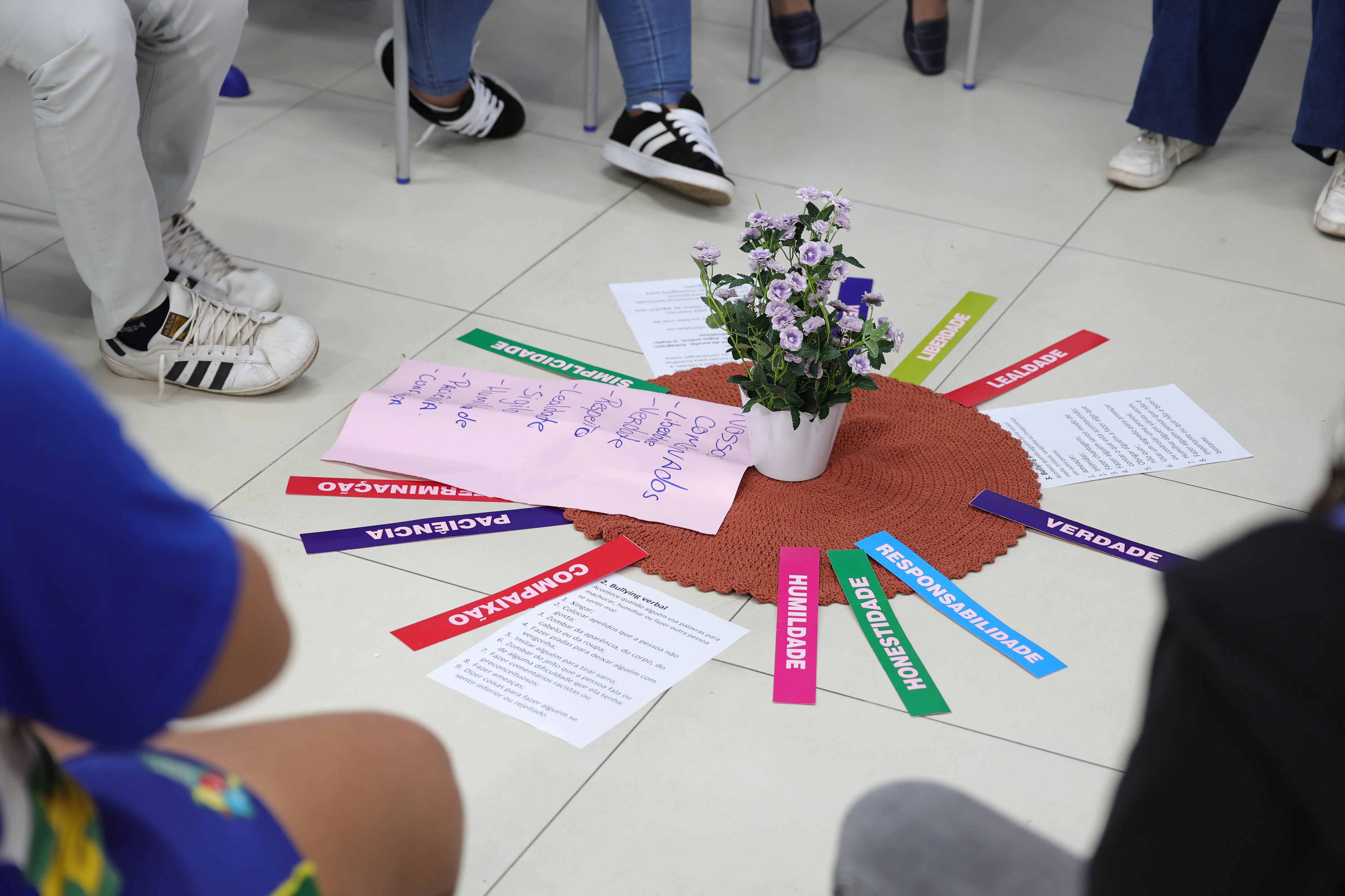 Plano detalhe do centro da roda de conversa no chão, mostrando um vaso de flores roxas em um tapete circular. Em volta, há papéis com textos e tiras coloridas com palavras como "RESPONSABILIDADE" e "COMPAIXÃO".