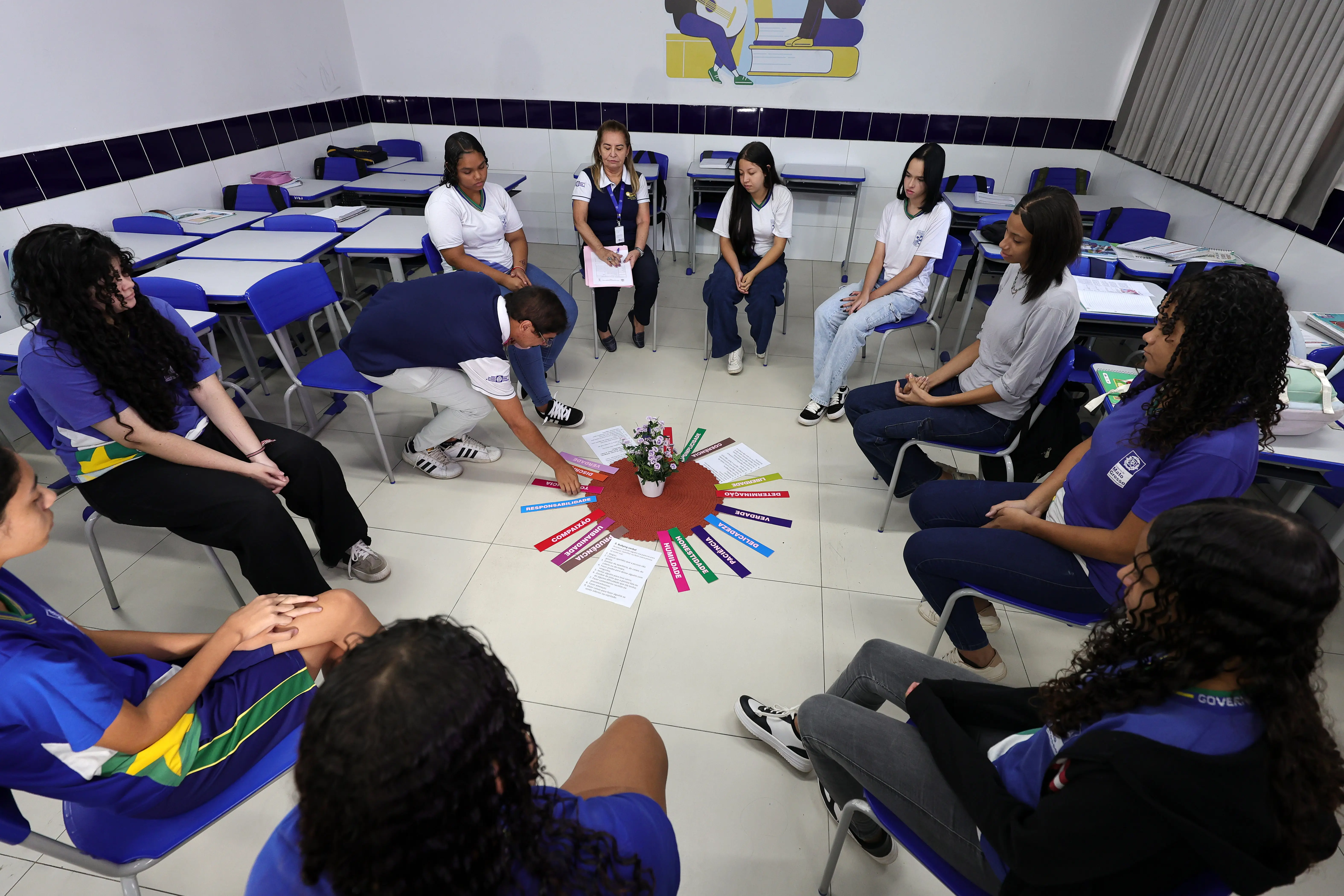 Estudantes e adultos dispostos em círculo em uma sala de aula. No centro do chão, há um vaso com flores sobre um tapete circular cercado por tiras coloridas com palavras escritas e folhas de papel.