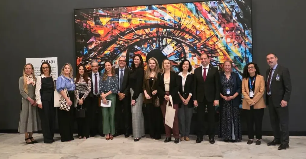 Quinze pessoas posam lado a lado diante de grande painel colorido com imagem de um olho, em espaço de piso claro. Alguns usam crachás, e dois homens de terno estão nas pontas.