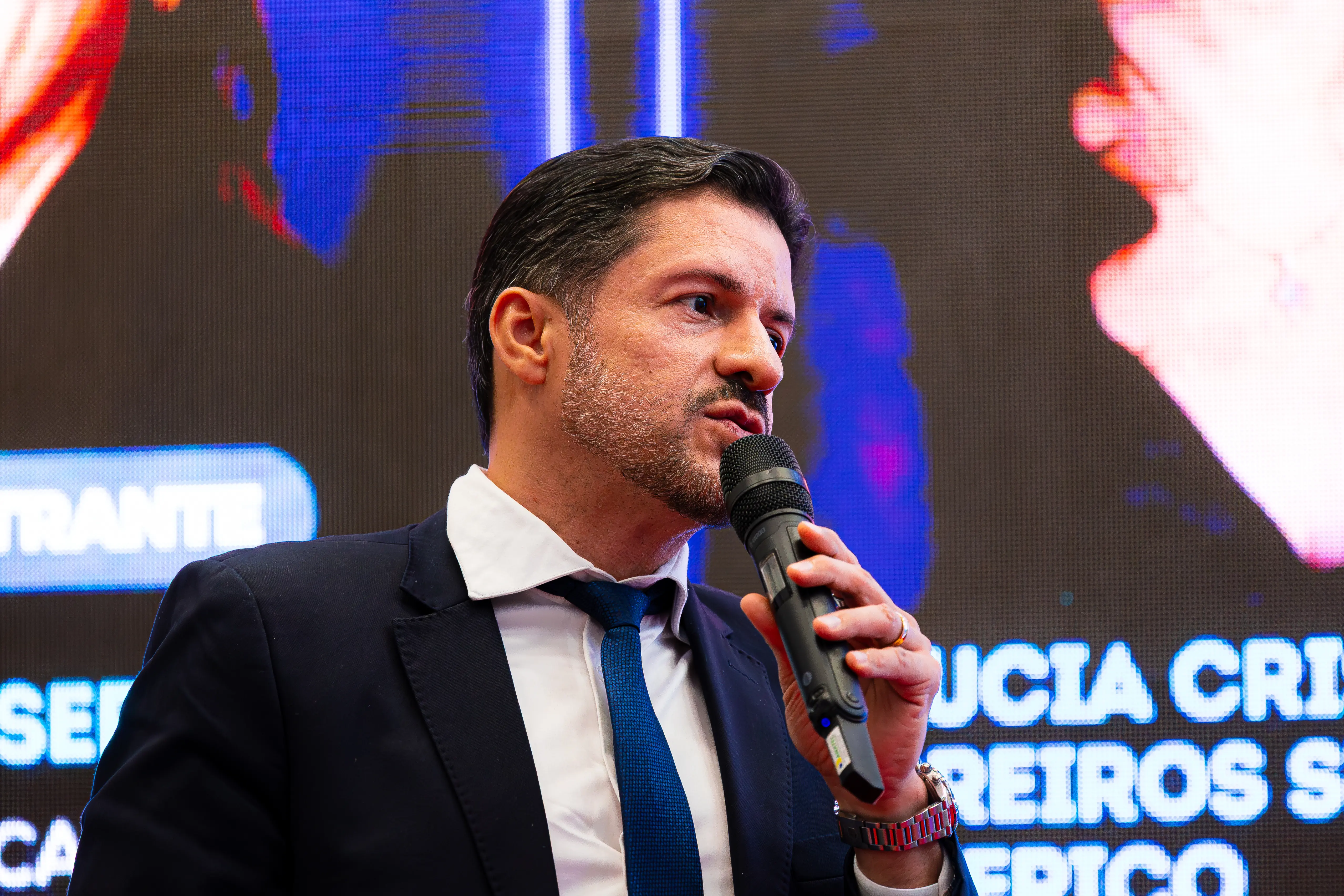 Homem de barba aparada, terno escuro e gravata azul discursa ao microfone.