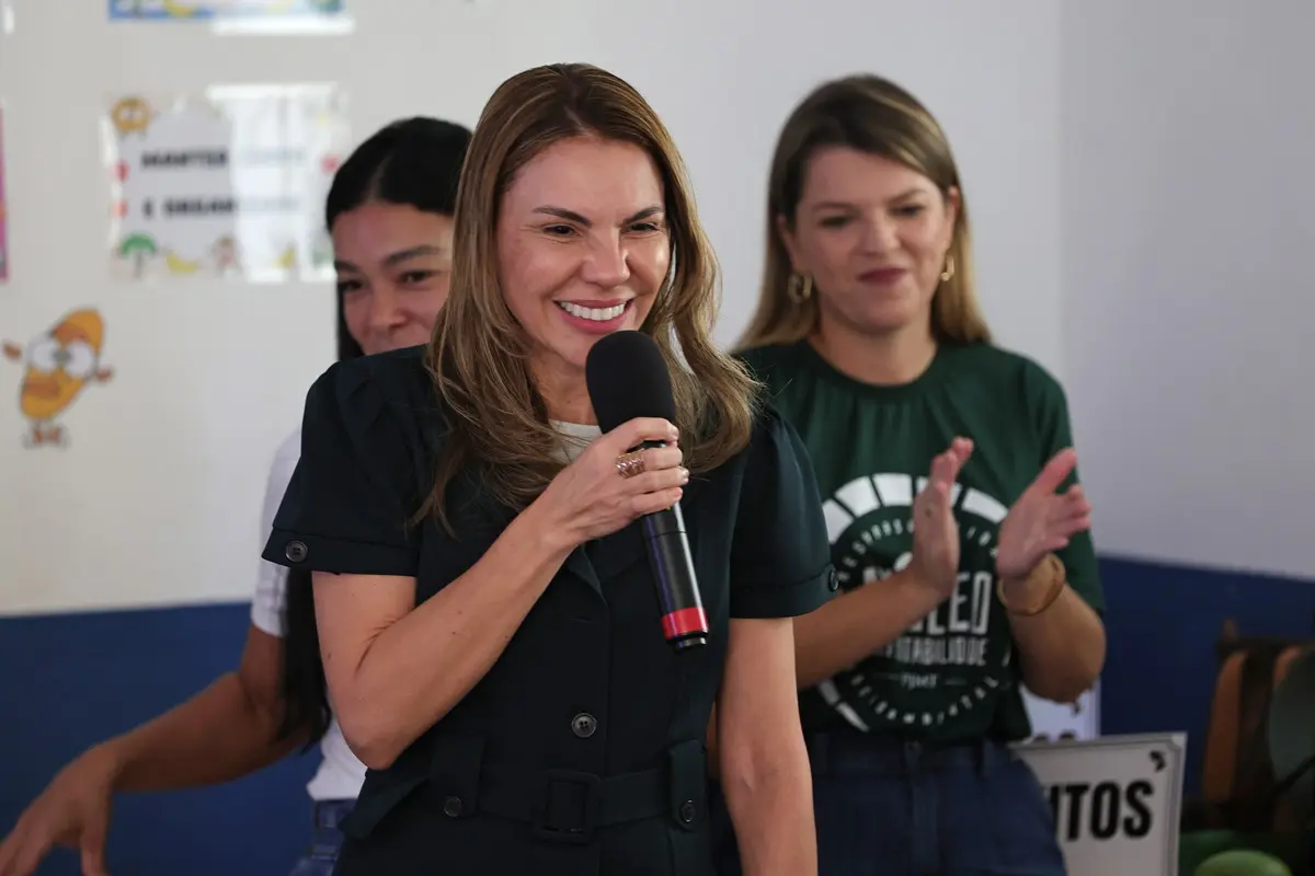 Mulher sorridente de cabelos castanhos fala ao microfone vestindo vestido preto. Ao fundo, outras mulheres acompanham a apresentação em sala com parede decorada.
