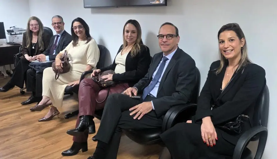 Três mulheres e dois homens de terno, sentados em cadeiras pretas, sorriem para a foto em sala de espera com piso de madeira. Uma delas segura bolsa ao colo; ao fundo, impressora e TV.