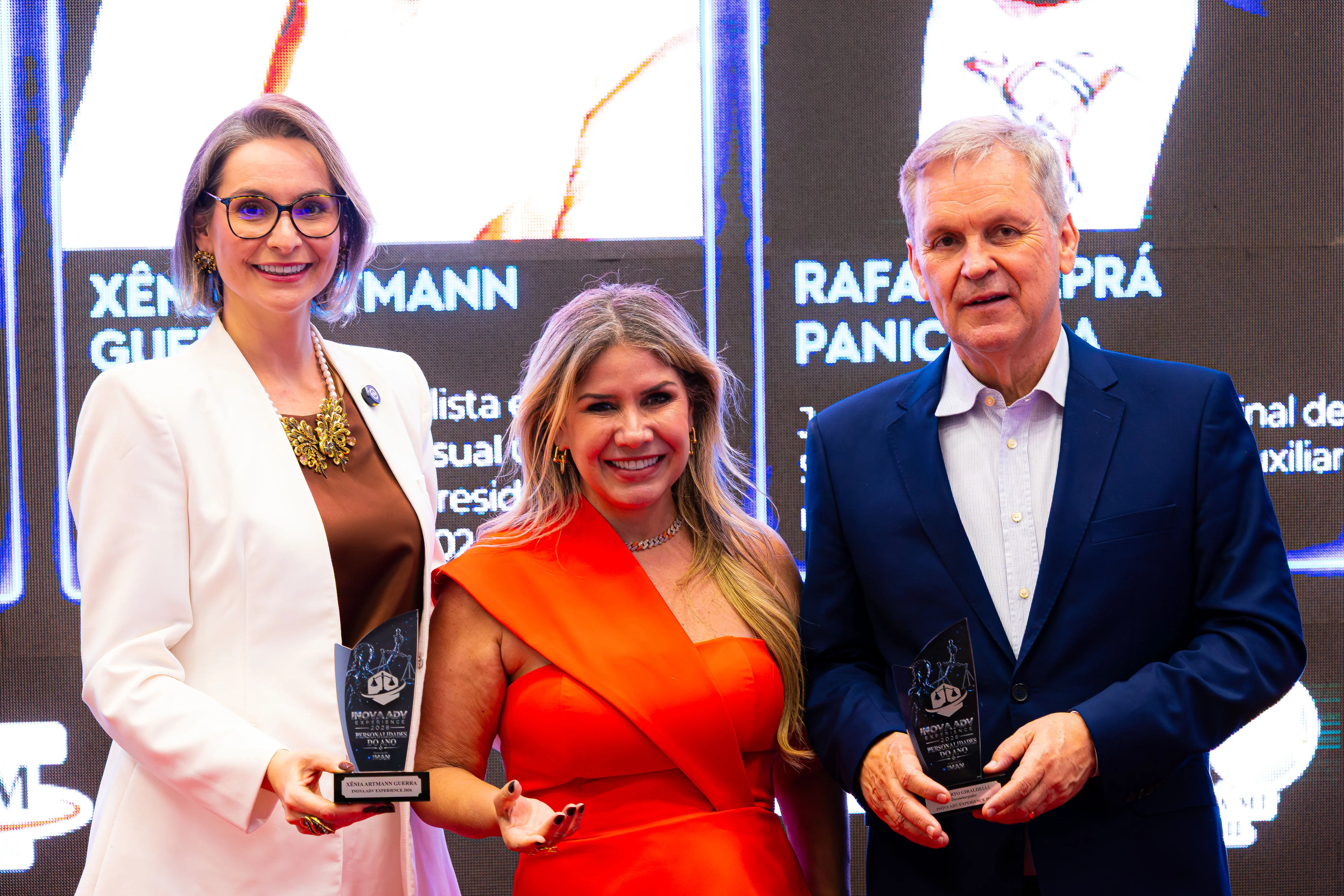 Duas mulheres e um homem sorriem para a foto segurando troféus. Ela usa blazer branco, a do meio vestido laranja e ele terno azul escuro, à frente de um painel de LED iluminado.