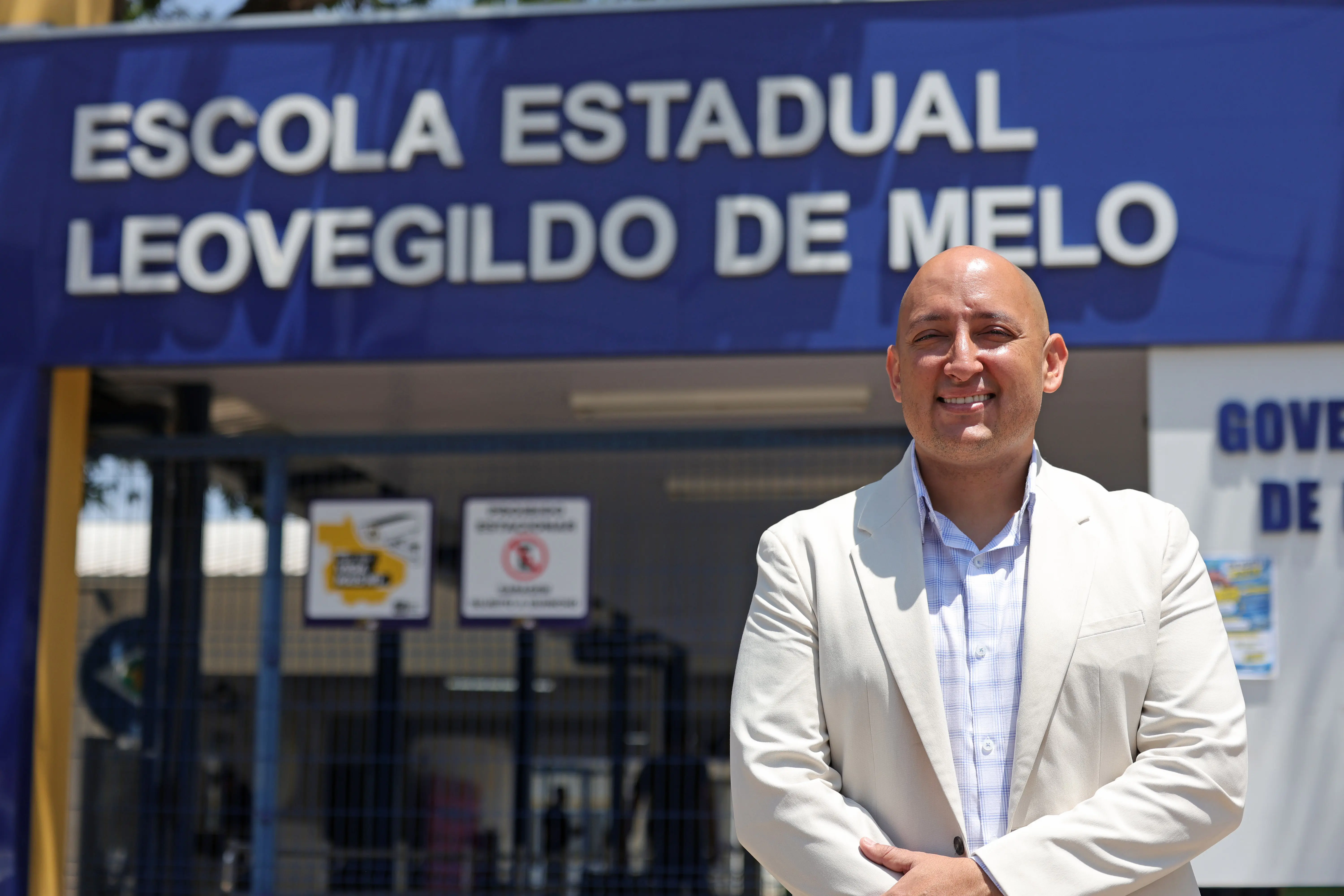 Um homem calvo, sorridente, vestindo blazer claro e camisa xadrez, em pé na frente de um prédio. Acima dele, há uma fachada azul com a inscrição em letras brancas: "ESCOLA ESTADUAL LEOVEGILDO DE MELO".