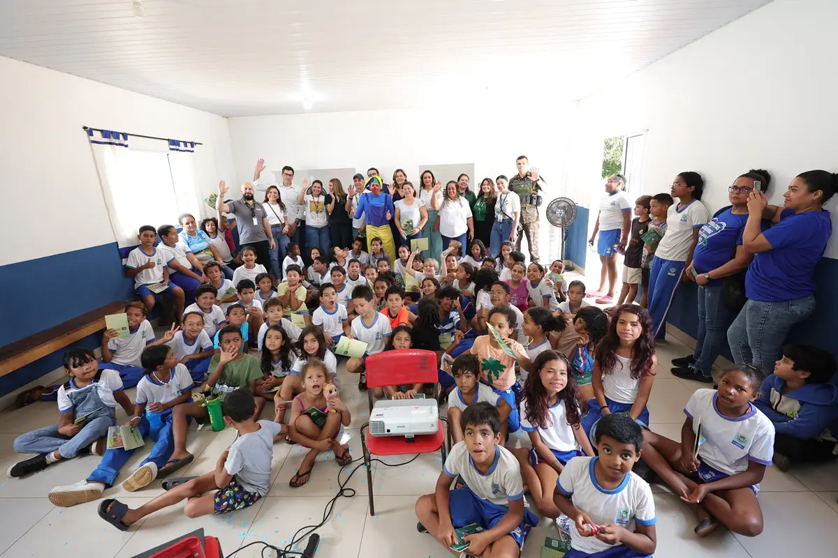 Crianças e adultos reunidos em sala de aula de paredes brancas e azuis. As crianças estão sentadas no chão e os adultos em pé ao fundo, com um projetor em destaque no centro.