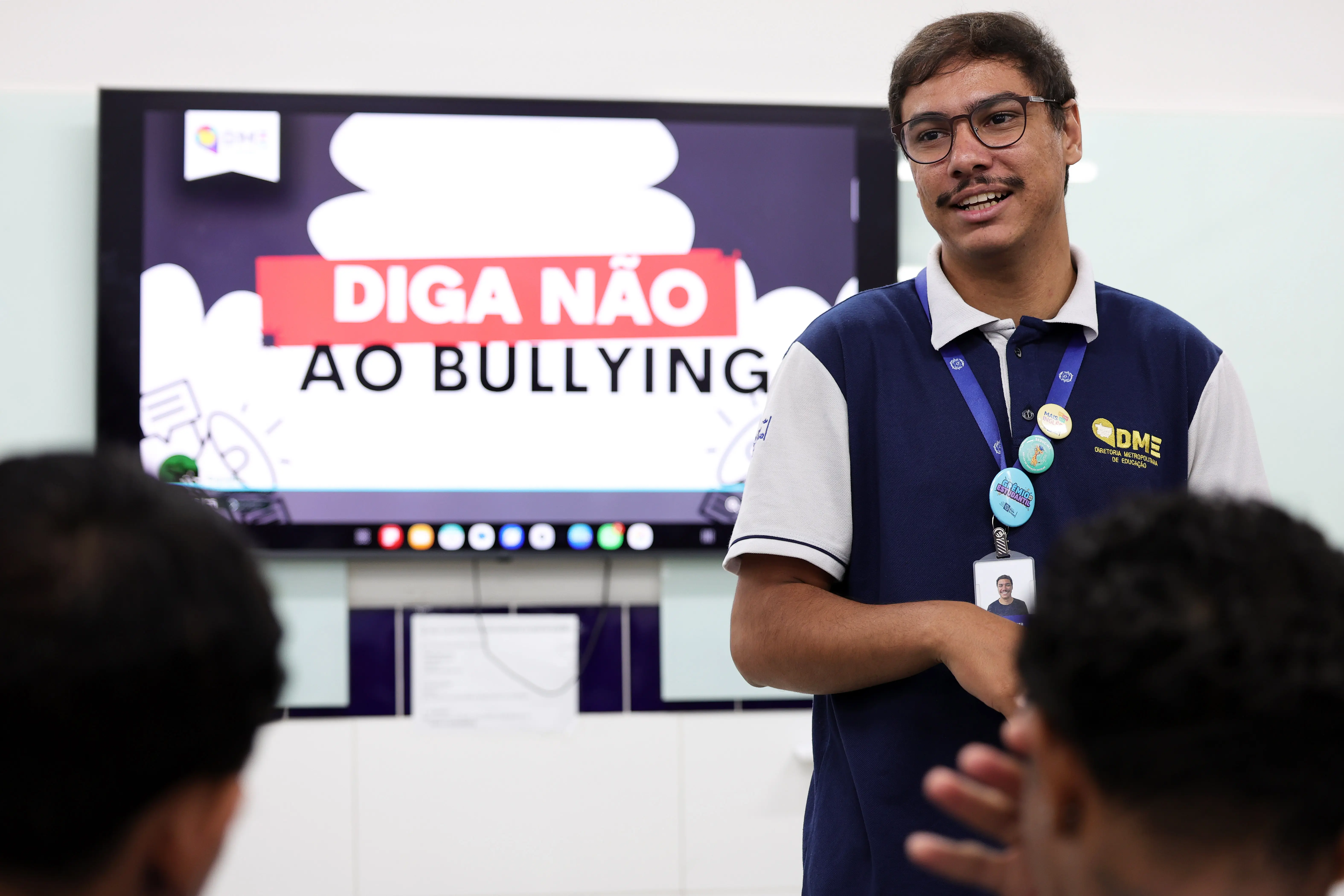 Um homem de óculos e camisa azul fala de pé em uma sala de aula diante de alunos sentados. Ao fundo, uma tela exibe "DIGA NÃO AO BULLYING" e duas pessoas observam em pé.