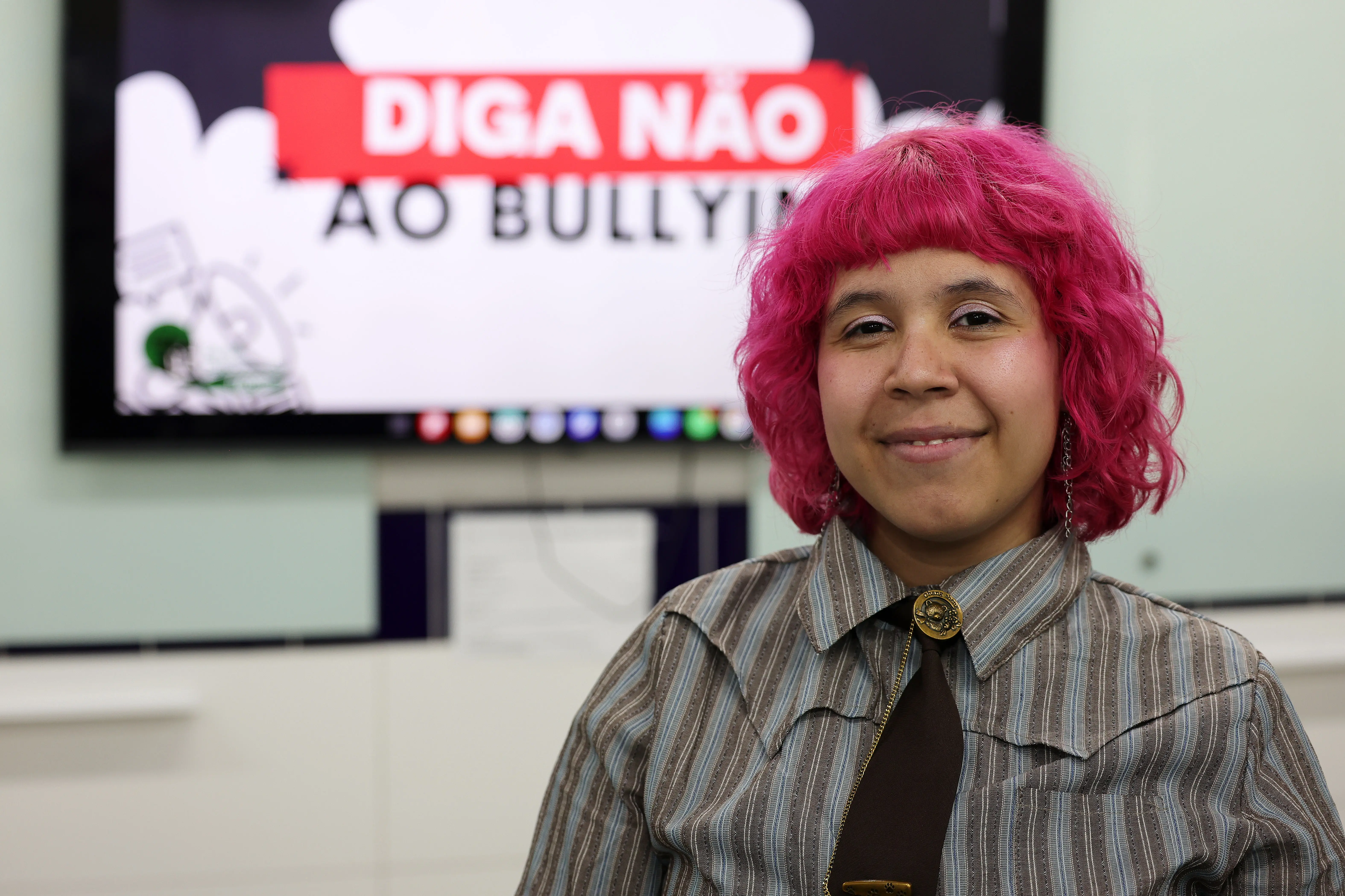 Retrato em plano médio de uma pessoa sorrindo, com cabelo rosa-choque, vestindo camisa listrada e gravata escura com broche. Ao fundo desfocado, vê-se a tela com o texto "DIGA NÃO AO BULLYING".