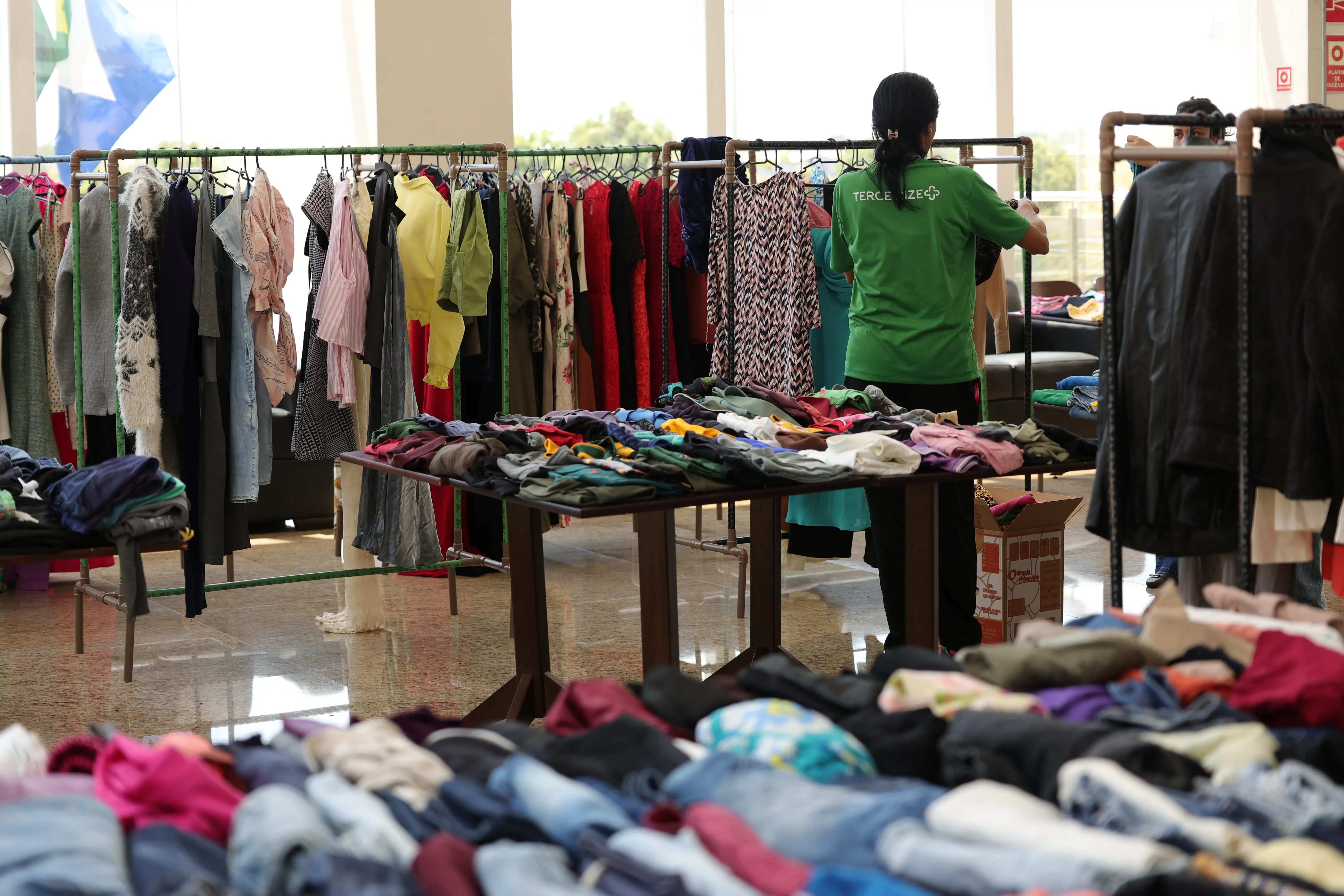 Fotografia de um ambiente de bazar de roupas. Mesas cobertas com peças de vestuário dobradas e araras repletas de cabides com roupas variadas estão dispostas no espaço. Ao fundo, janelas grandes e uma pessoa de costas com camiseta verde.