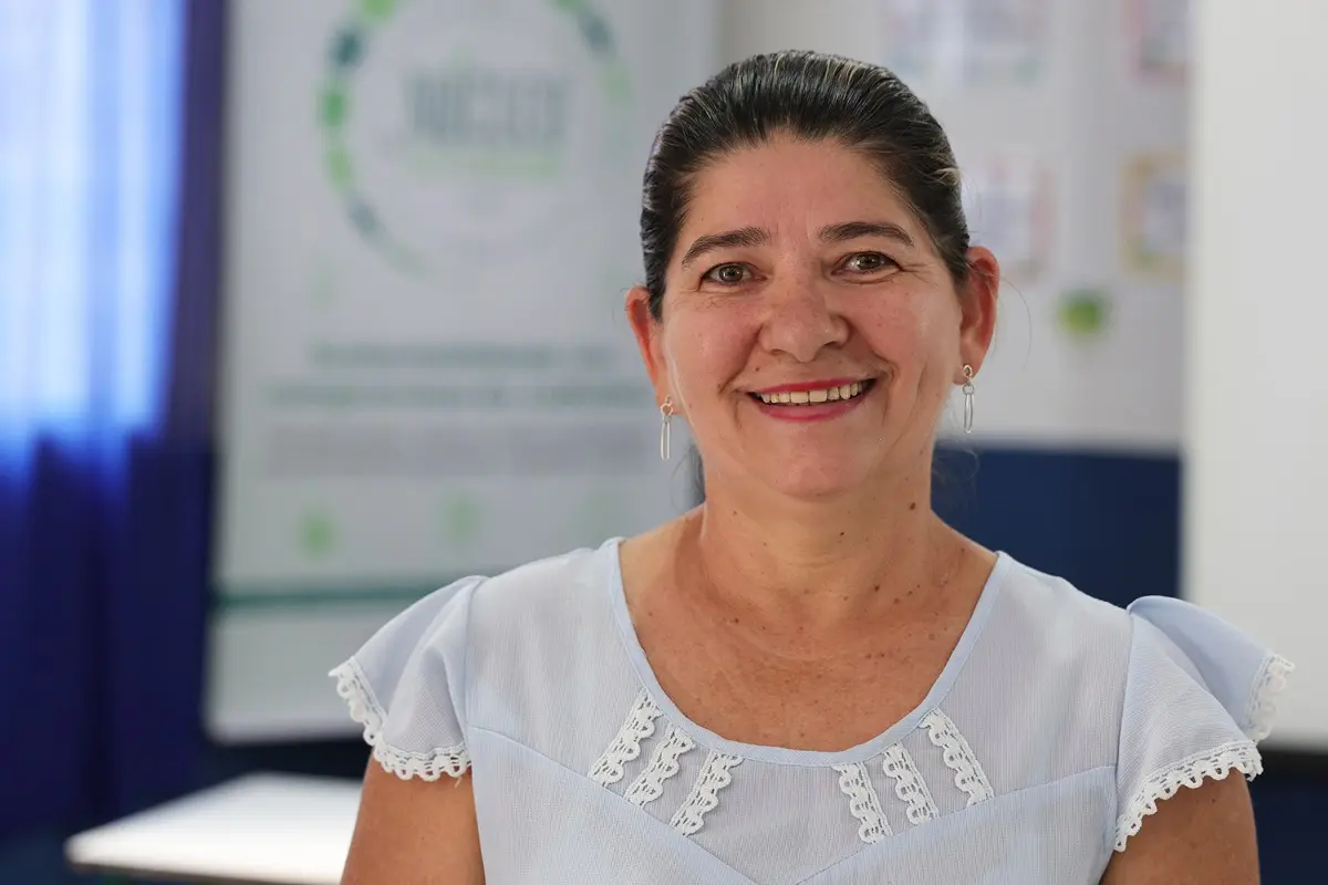 Mulher de cabelos escuros presos sorri para a câmera. Usa blusa azul clara com detalhes em renda. Ao fundo desfocado, há um banner institucional com logomarca circular.