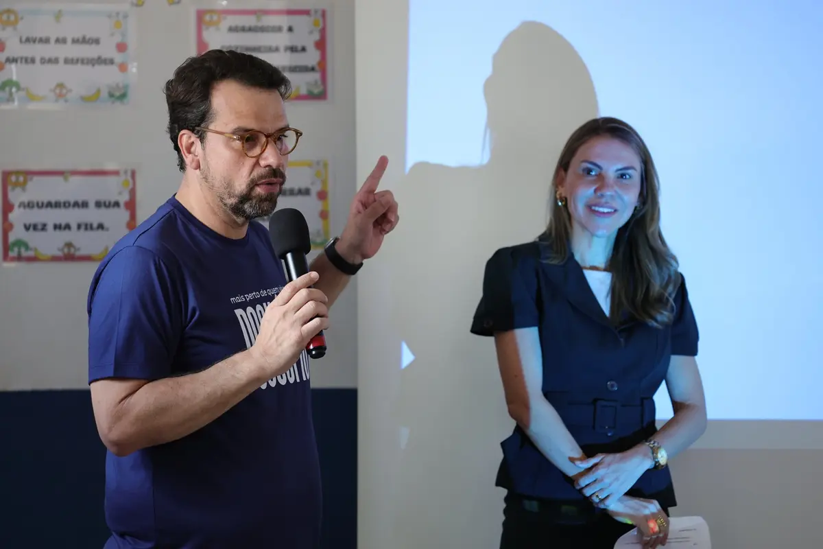 Homem de óculos fala ao microfone e gesticula com o indicador erguido, vestindo camiseta azul escura. Ao lado, mulher de vestido escuro escuta sorrindo. Ao fundo, parede com cartazes.