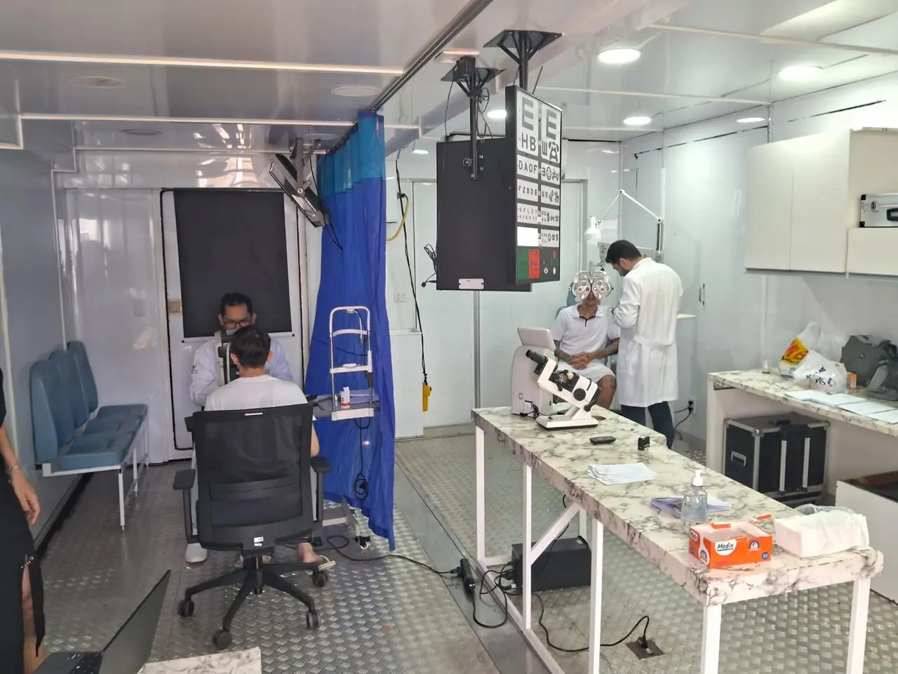 Consultório oftalmológico móvel moderno em trailer, com profissionais de jaleco branco realizando exames de vista em dois pacientes. Há equipamentos clínicos, tabela Snellen e divisória azul.