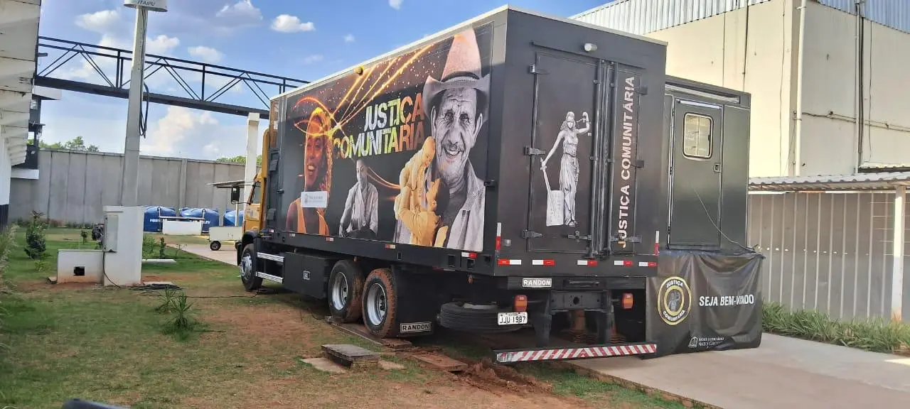 Caminhão do projeto "Justiça Comunitária" estacionado ao ar livre. O baú preto traz adesivo com o nome do programa, fotos de pessoas e o símbolo da Justiça na traseira.