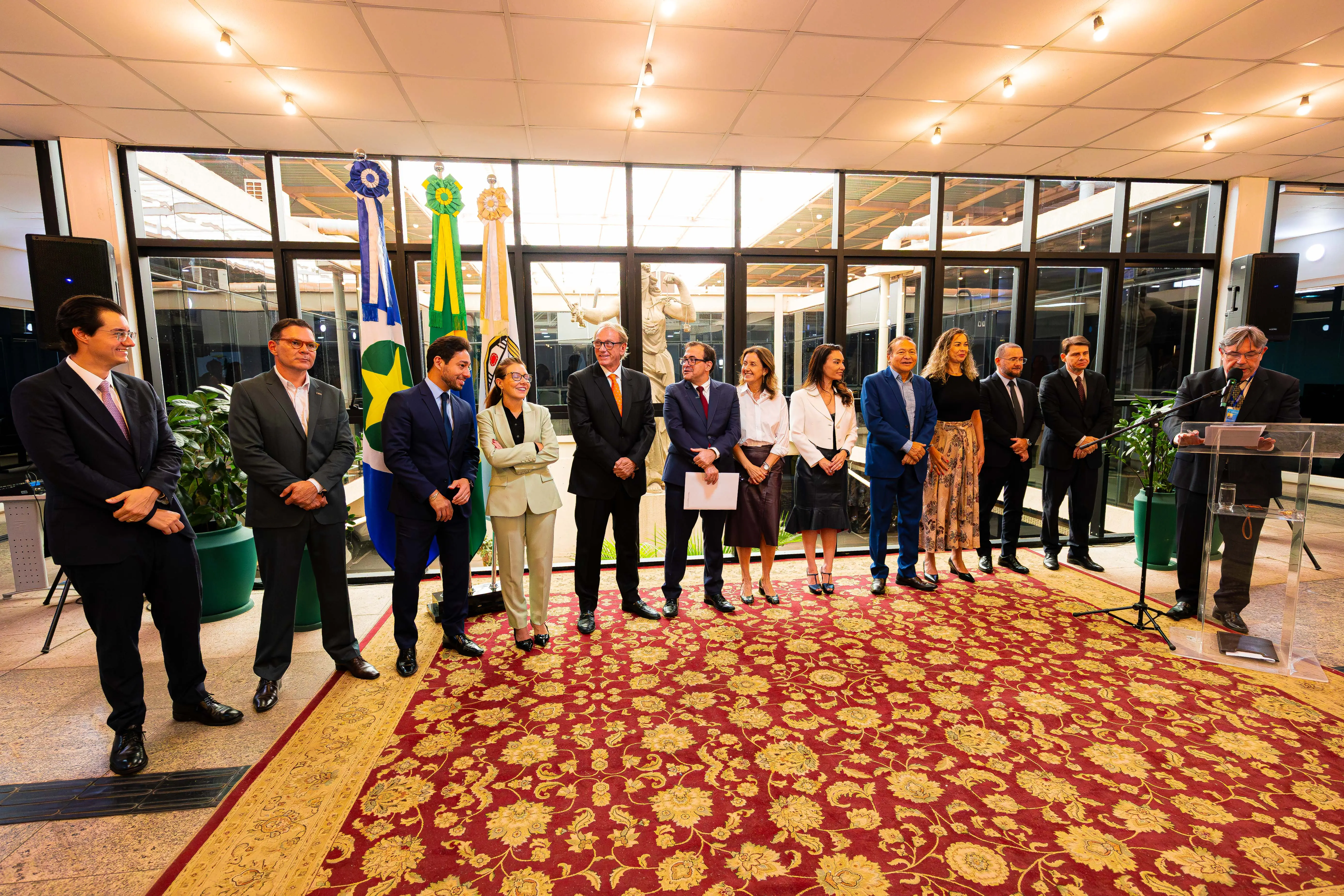 Grupo em traje formal alinhado sobre tapete vermelho floral em salão iluminado. À direita, homem discursa em púlpito transparente. Ao fundo, bandeiras oficiais e parede de vidro.