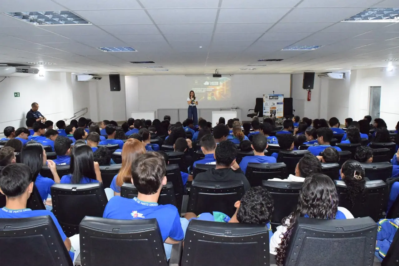 Mulher discursa no palco de auditório lotado por estudantes de camiseta azul. Vista traseira da plateia sentada em poltronas pretas, voltada para a palestrante e a projeção ao fundo.
