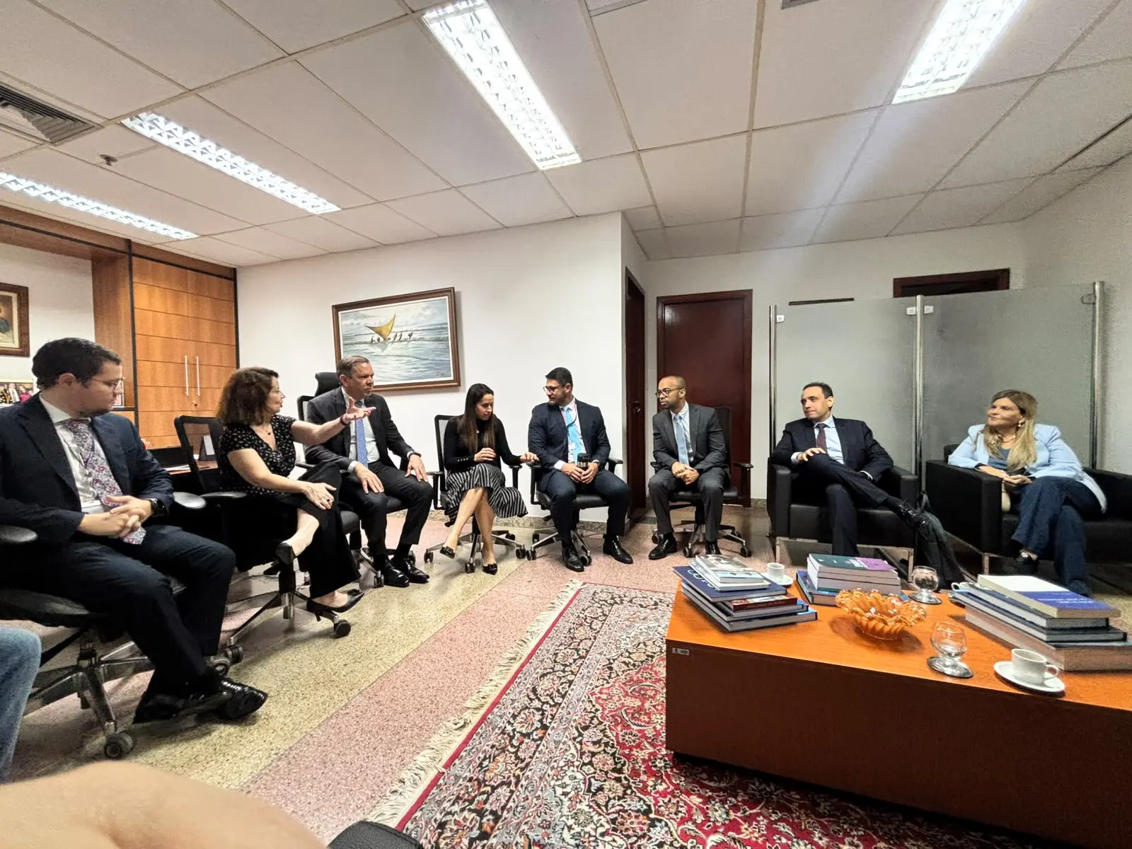 Grupo de pessoas reunido em um escritório conversando acomodadas em cadeiras e poltronas dispostas em círculo. Uma mulher gesticula enquanto fala para os demais participantes da reunião. No chão, destaca-se um tapete estampado e uma mesa de centro repleta de livros. 