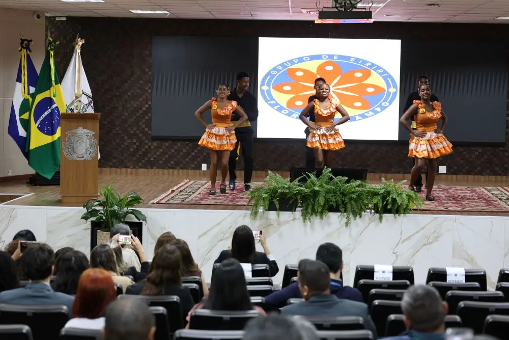 Foto horizontal colorida, em plano aberto, que mostra três casais de bailarinos dançando siriri no palco do auditório do TJMT, de frente para a plateia. No telão, aparece a logomarca do Grupo de Siriri e Cururu Flor do Campo.