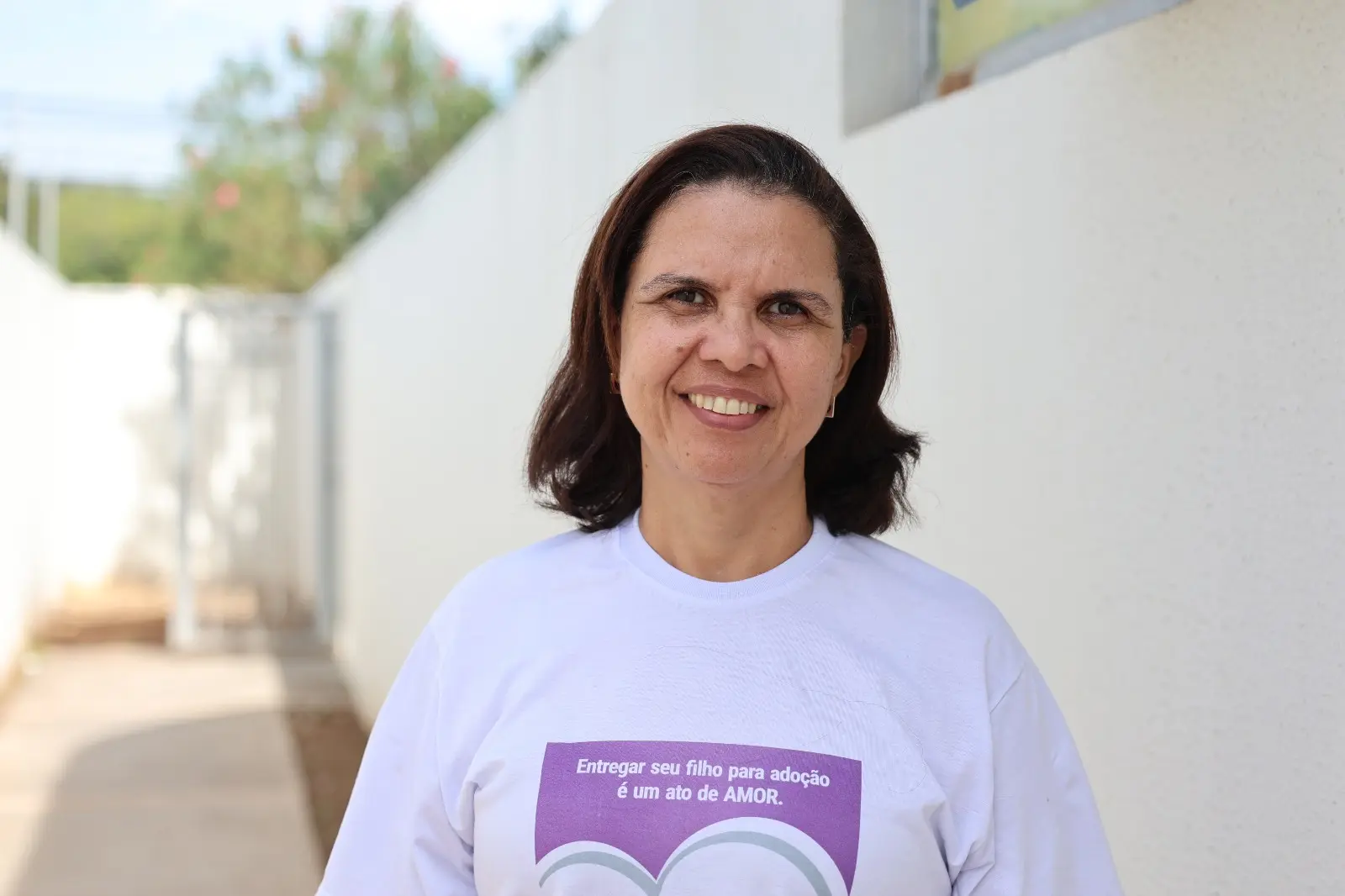A imagem mostra a assistente social da Vara da Infância e Juventude de Cuiabá, Cedirlene Cunha, posando para foto em área externa. Ela veste camiseta branca da campanha Entrega Legal, com a frase 