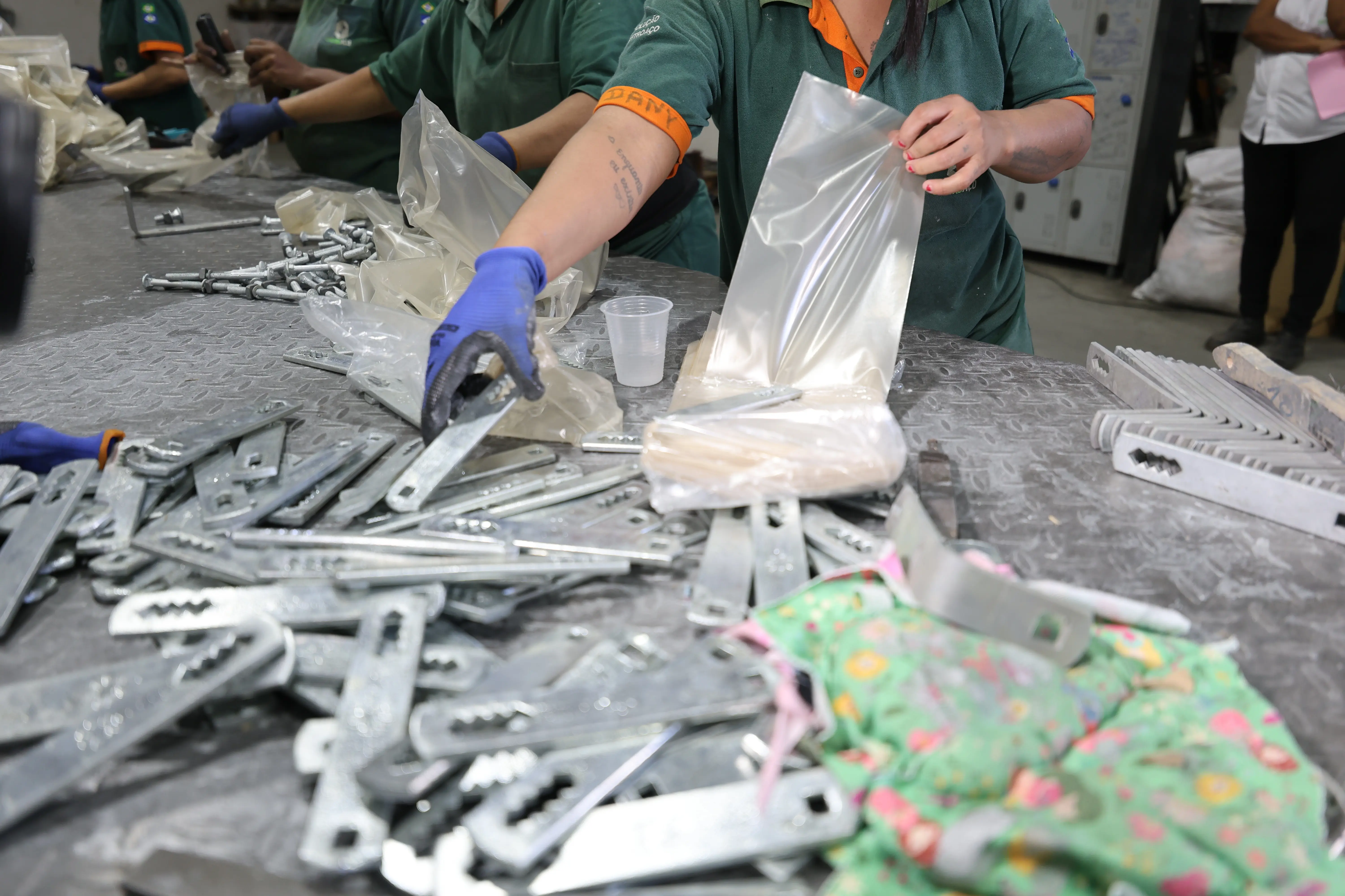 Reeducanda de uniforma verde e luva azul segura um plástico com uma mão e manuseia uma peça metálica com a outra.