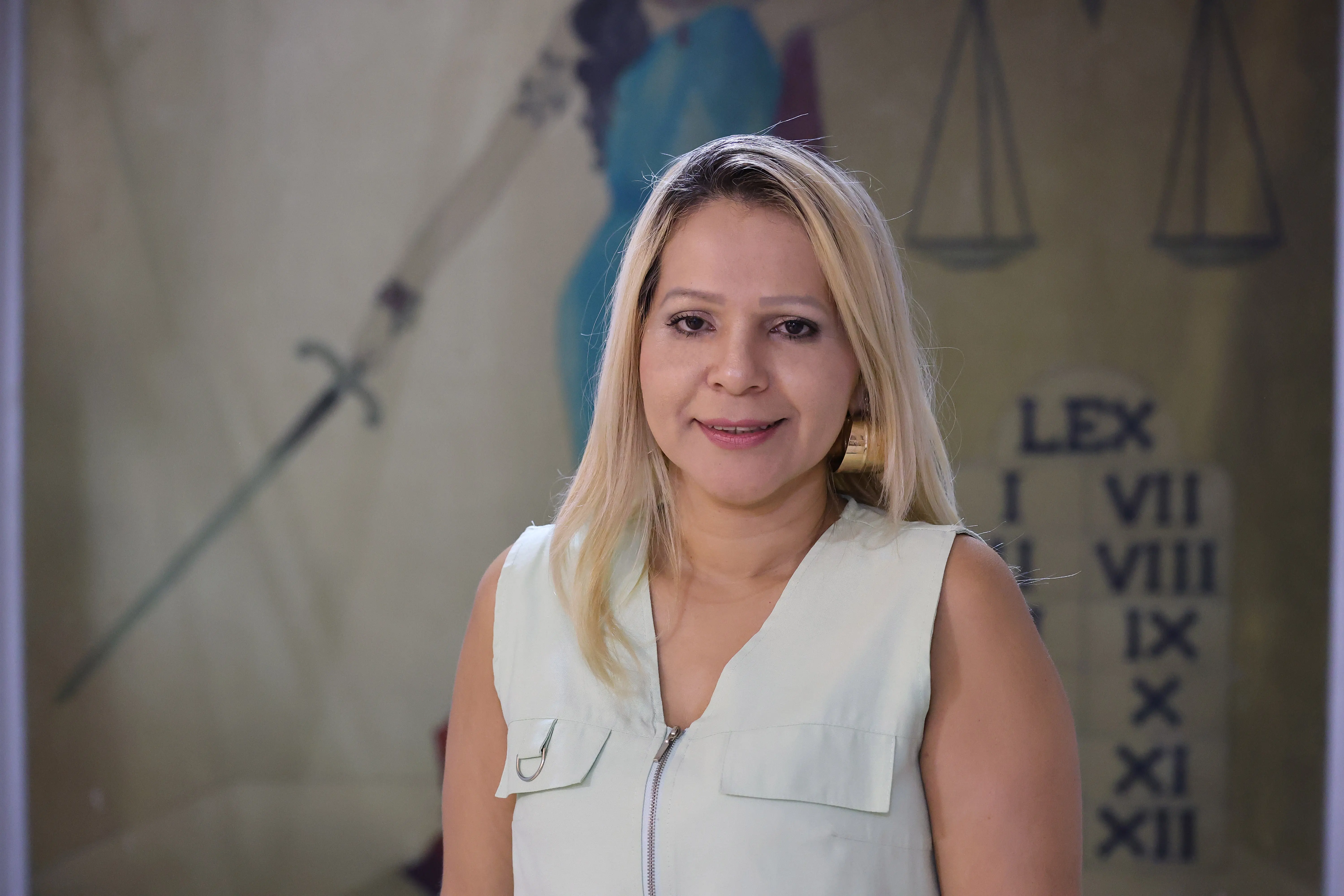 Retrato de Arlene, uma mulher loira sorrindo com blusa clara sem mangas. Ao fundo, uma imagem estilizada da deusa da justiça com uma espada e a balança.