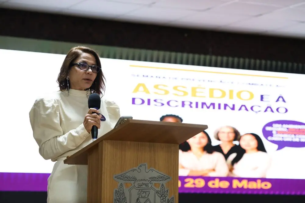 Foto horizontal colorida que mostra a desembargadora Juanita Clait Duarte falando ao microfone, no púlpito do auditório do TJMT. Ela é uma senhora de pele negra, cabelos castanhos na altura dos ombros, usando calça e blusa de manga longa bege. Atrás dela, há um grande telão com a logomarca do evento