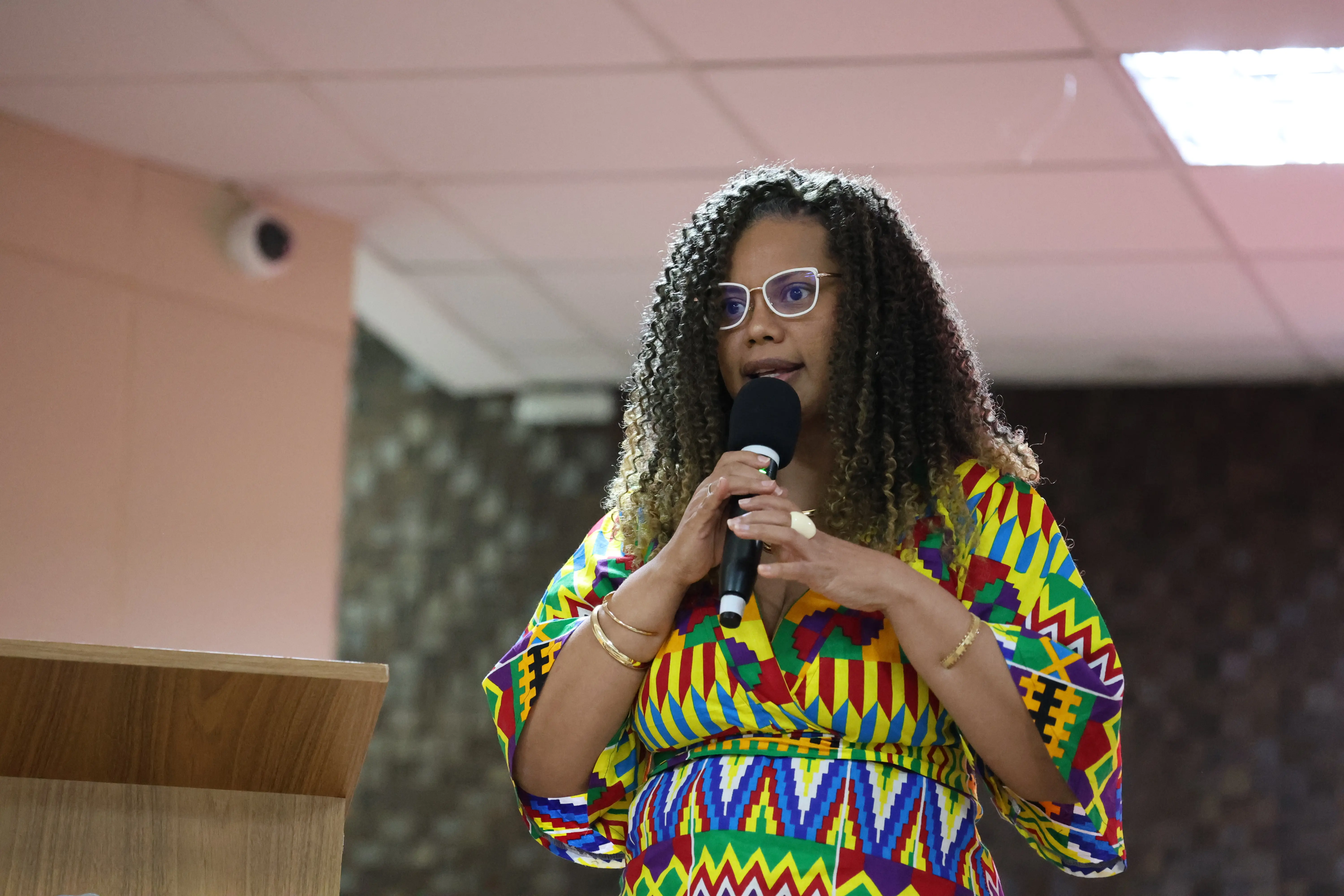 Foto da professora Silviane, uma mulher negra de cabelos cacheados e óculos, falando ao microfone no palco. Ela veste uma roupa com estampa geométrica colorida.