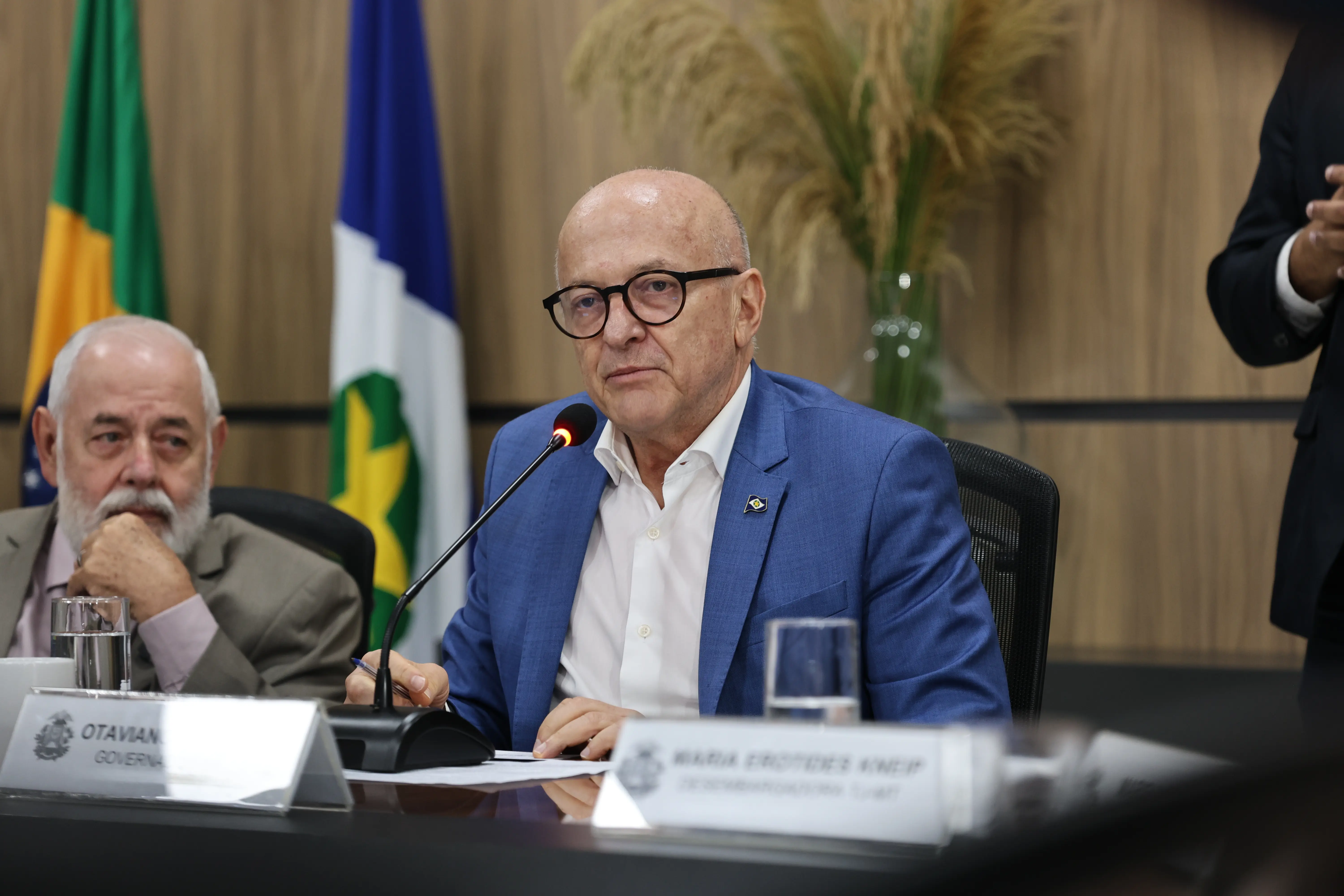 Homem careca de óculos pretos e terno azul fala ao microfone em uma mesa de reuniões. À esquerda, outro homem idoso de barba branca observa com a mão no queixo. Ao fundo, há bandeiras oficiais e uma parede amadeirada.