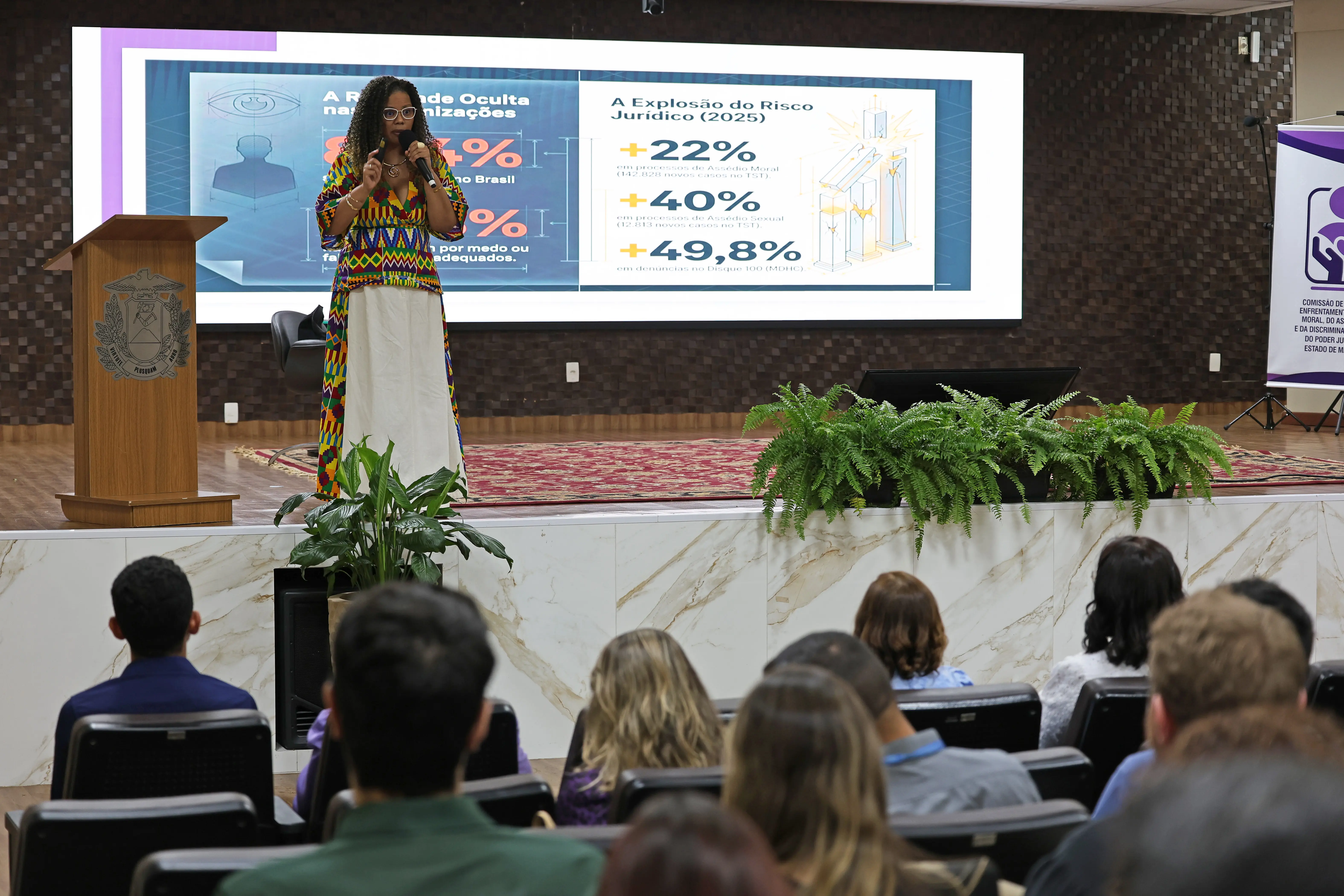 Mulher negra palestrando no palco com vestido colorido e saia branca. Ao fundo, um telão exibe dados sobre risco jurídico e assédio. Em primeiro plano, a plateia assiste de costas.