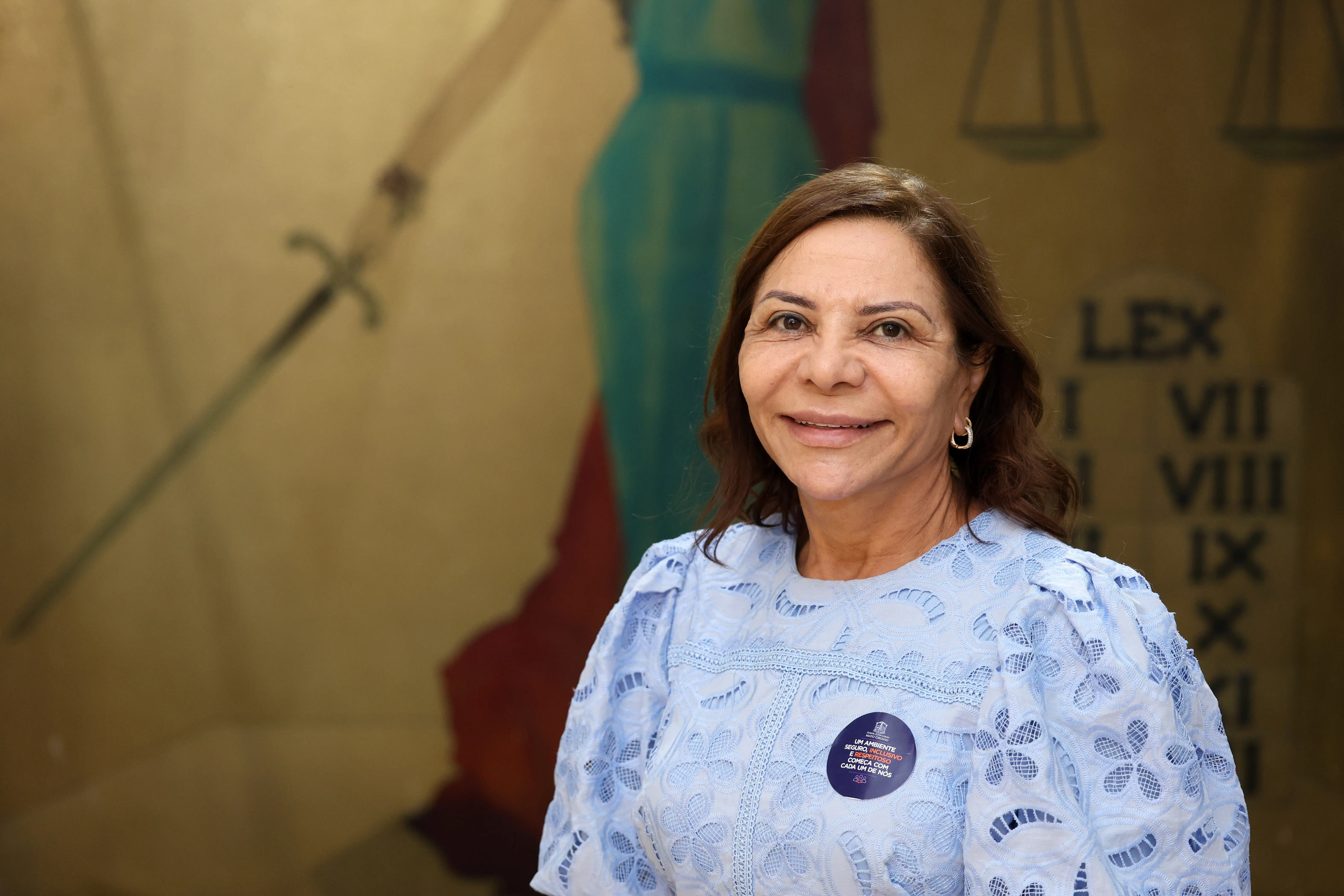 Retrato da desembargadora Juanita, uma mulher de cabelos castanhos sorrindo com blusa de renda azul claro e um bóton fixado na roupa.