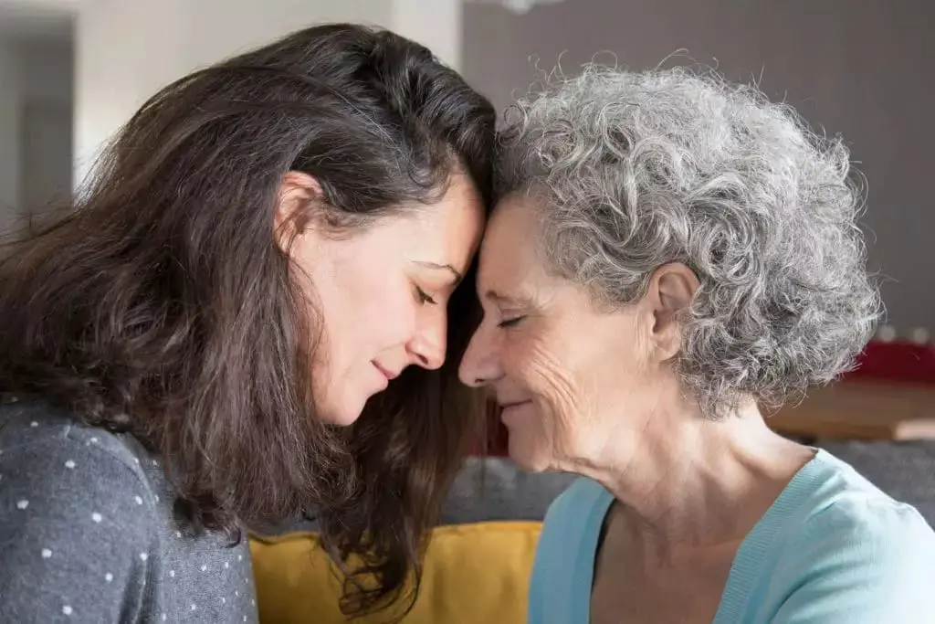 Foto colorida horizontal onde aparecem duas mulheres, uma jovem e uma idosa, encostando a testa uma na outra e sorrindo. A mulher da esquerda tem cabelos castanhos e volumosos e a mulher da direita tem cabelos cacheados grisalhos. Ao fundo há um cenário doméstico. 