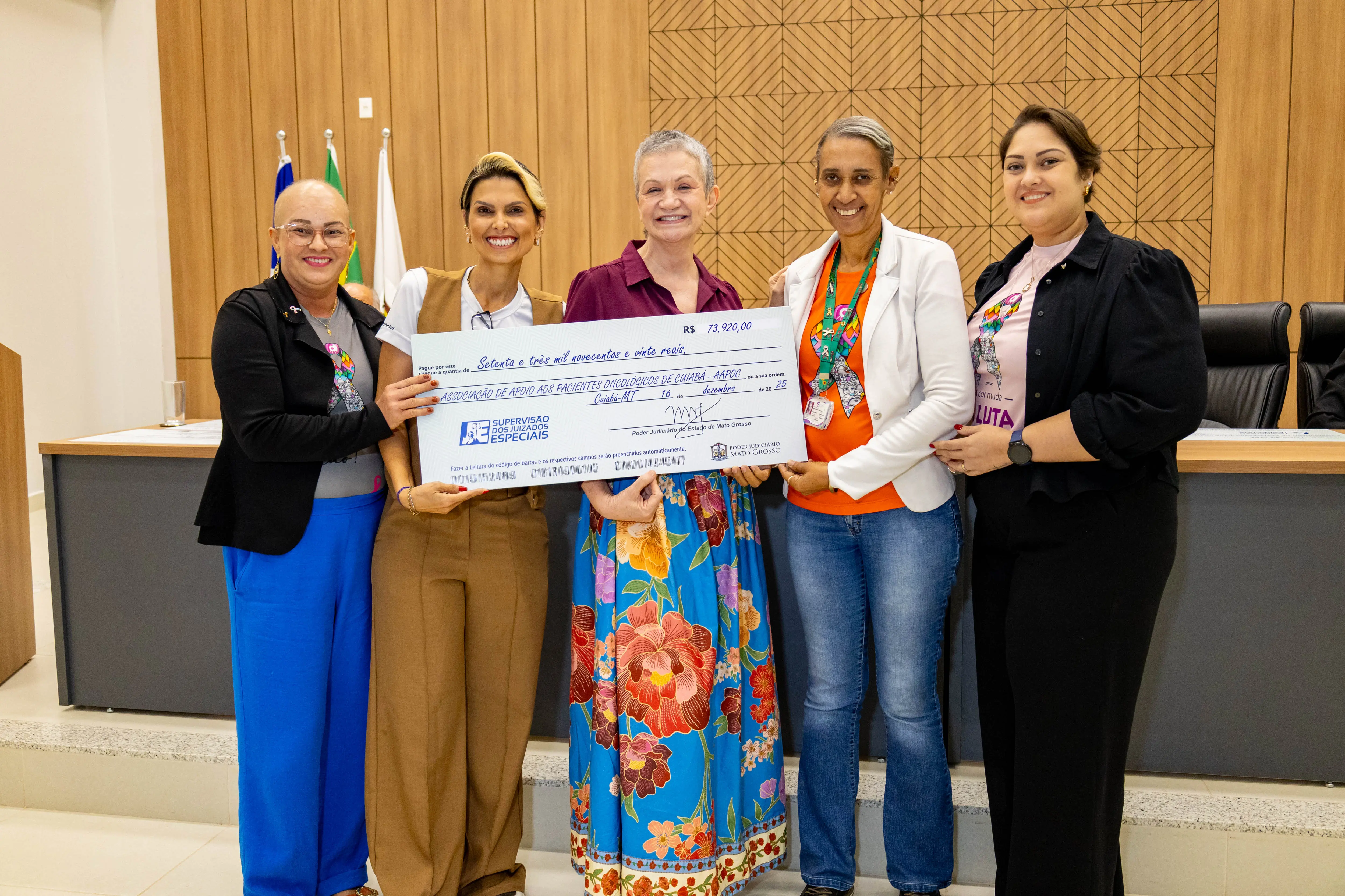 Cinco mulheres posam sorridentes segurando cheque simbólico. Vestem roupas sociais e coloridas. Ao fundo, mesa e bandeiras oficiais, evidenciando ato formal de destinação de recursos a entidade social.
