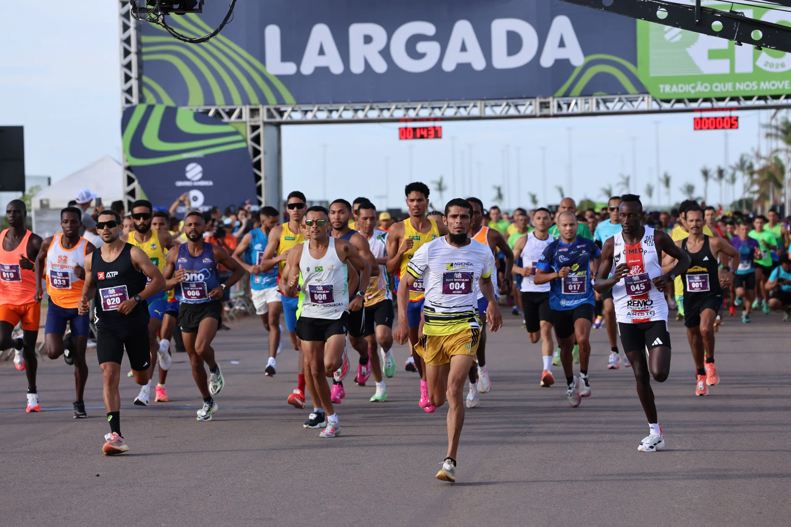 Multidão de corredores avança junta pela pista logo após a largada. Pessoas de diferentes idades ocupam todo o espaço, com sinalização e estrutura do evento visíveis.